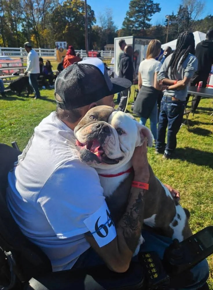 A man wearing a white shirt and a black cap hugging a large smiling bulldog at an outdoor event with people and tents in the background.