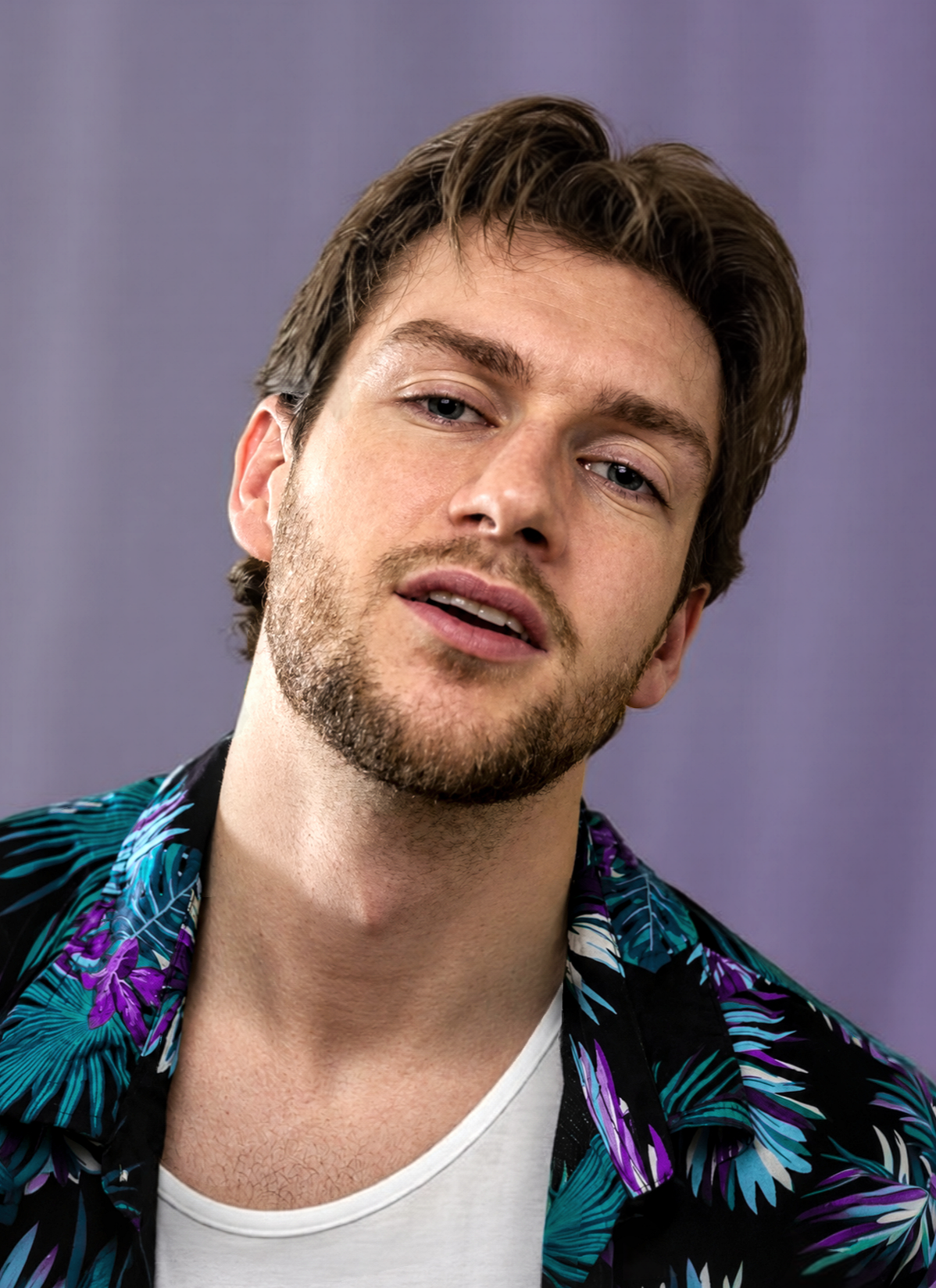 Lyall Hardy Grey, a young man with brown hair and a beard wearing a colorful Hawaiian shirt and a white t-shirt against a purple background.