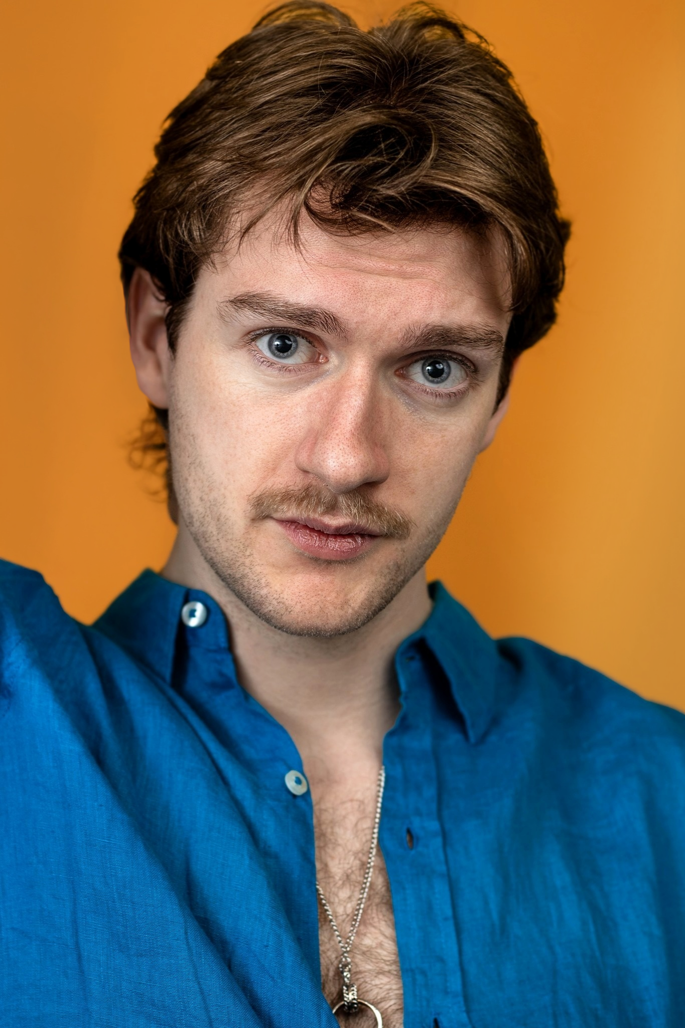 Close-up of Lyall Hardy Grey, a man with light skin, brown hair, and blue eyes, wearing a blue shirt with button undone, revealing chest hair and a silver necklace, against an orange background.