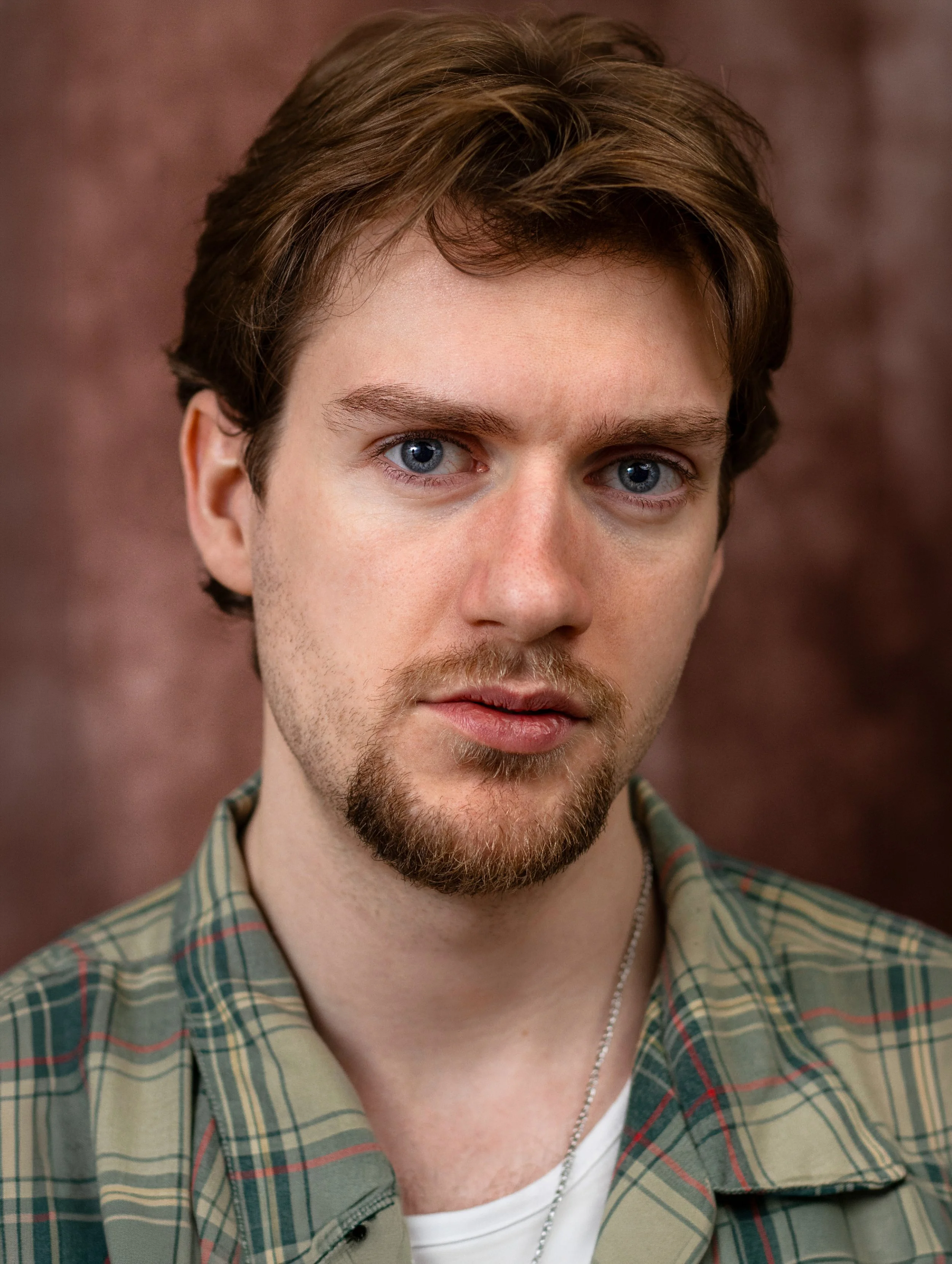 Close-up of Lyall Hardy Grey. A young man with brown hair, blue eyes, and a light beard, wearing a plaid shirt over a white T-shirt, with a silver chain necklace, against a blurred brown background.