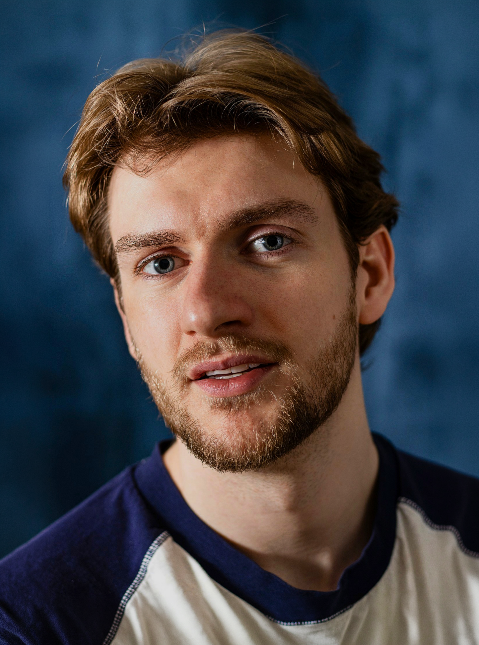 Close-up of Lyall Hardy Grey with blue eyes, brown hair, and light facial hair, wearing a navy and white sweatshirt, against a dark background.