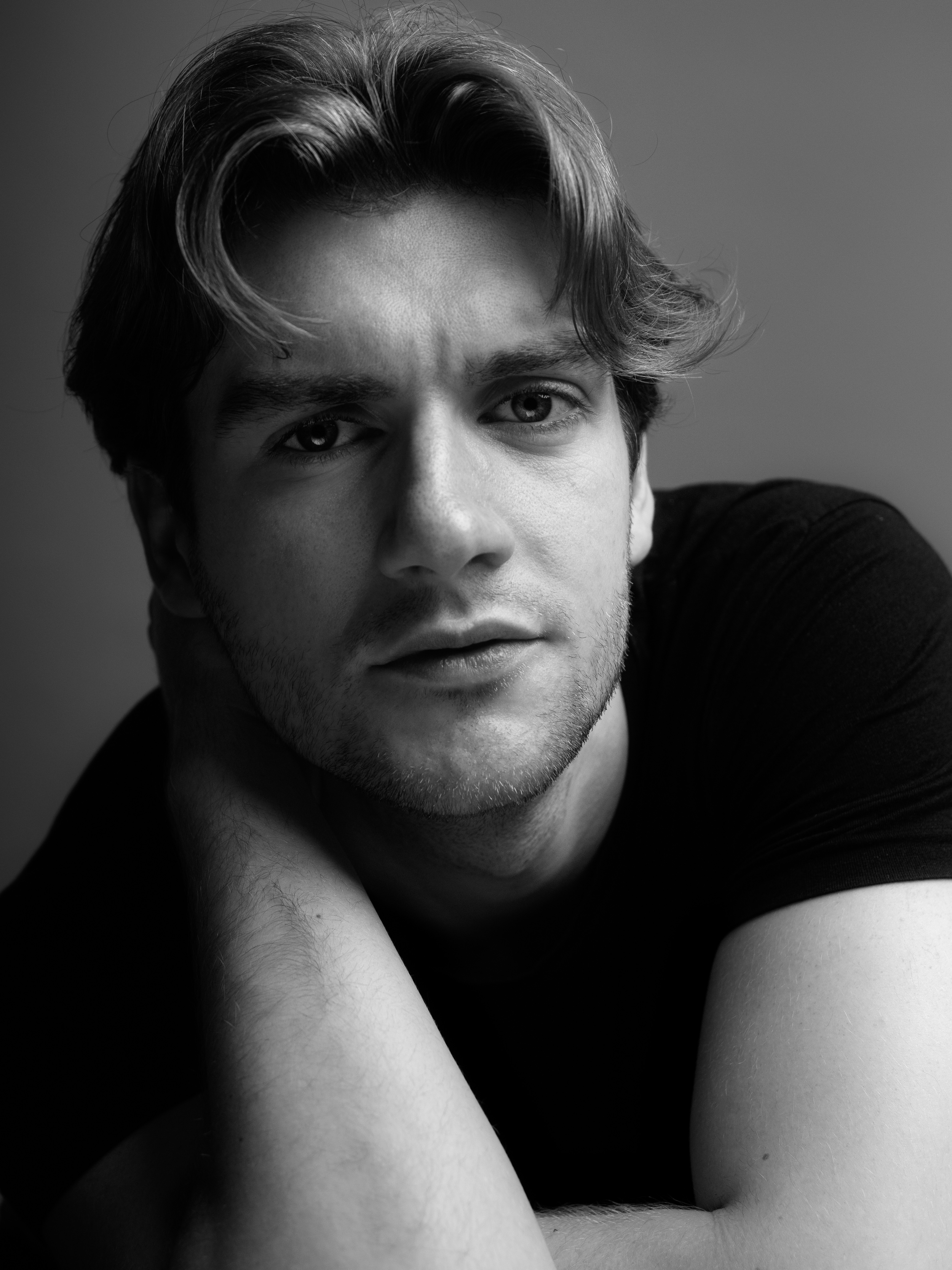 Black and white portrait of a man, Lyall Hardy Grey, with wavy hair, looking at the camera with a relaxed expression, resting his face on his crossed arms.