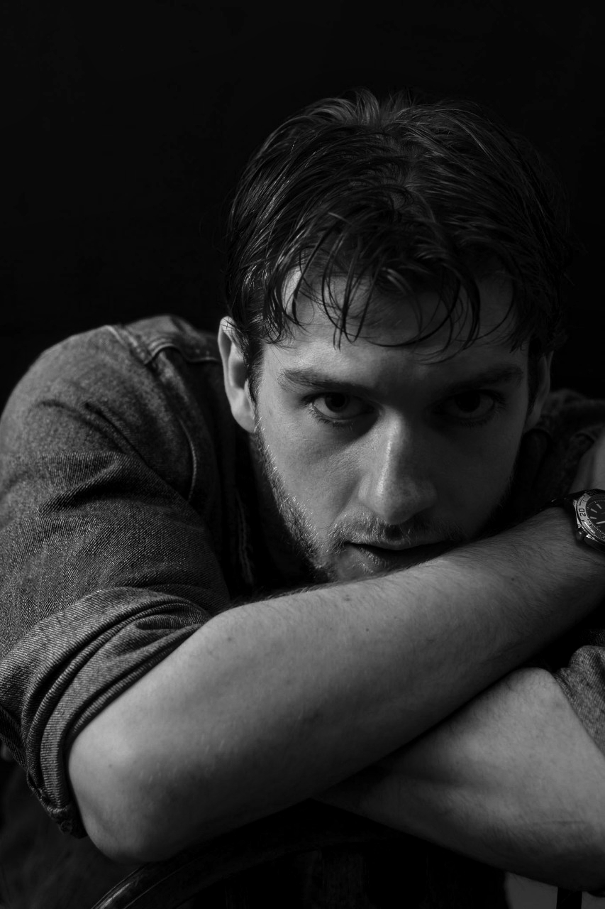 Black and white portrait of Lyall Hardy Grey. A young man with dark hair, leaning forward with arms crossed on a surface, staring intently at the camera.