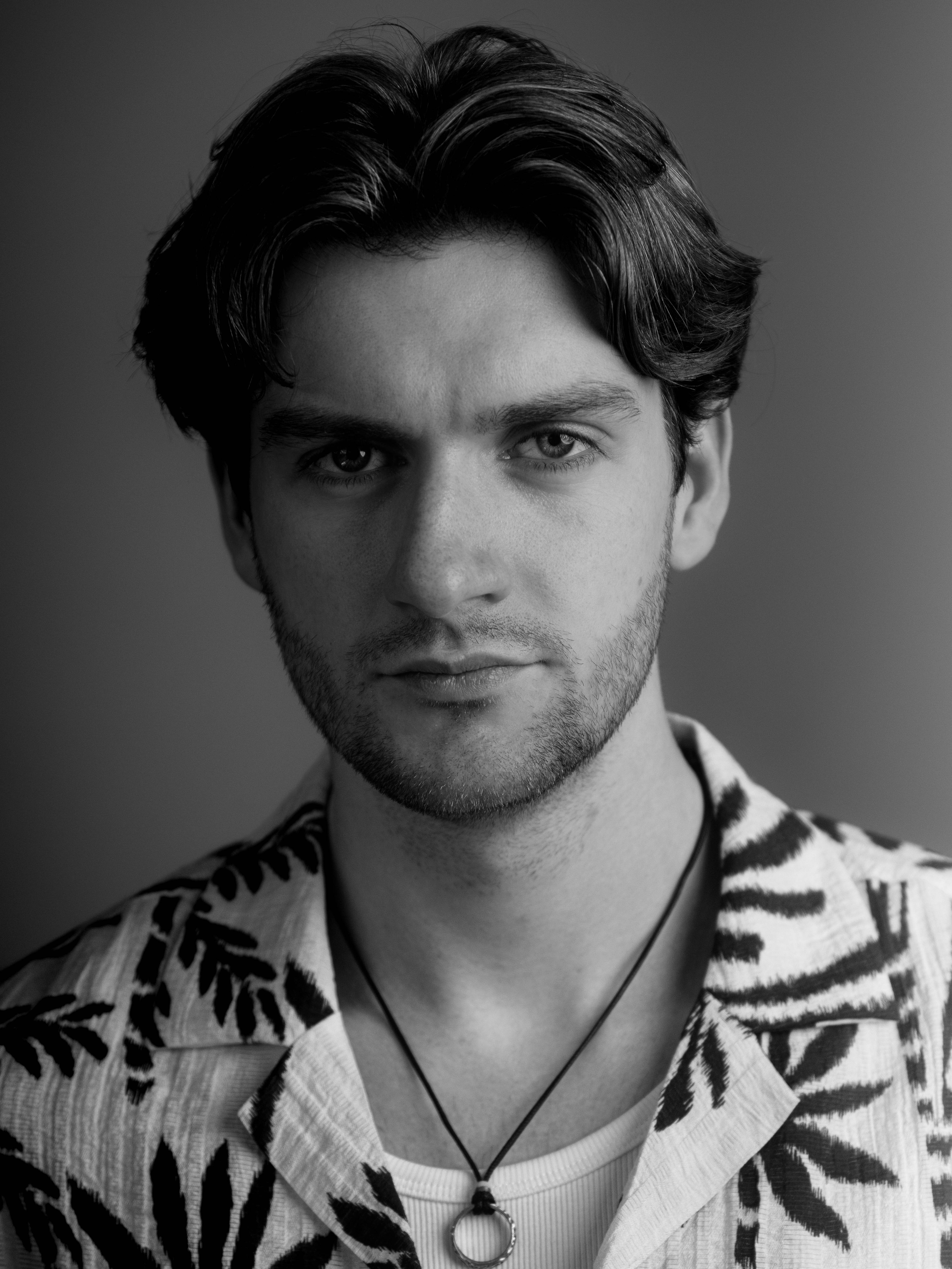 Black and white portrait of Lyall Hardy Grey, a young man with dark hair, wearing a striped shirt with animal print accents, and a necklace with a ring pendant.