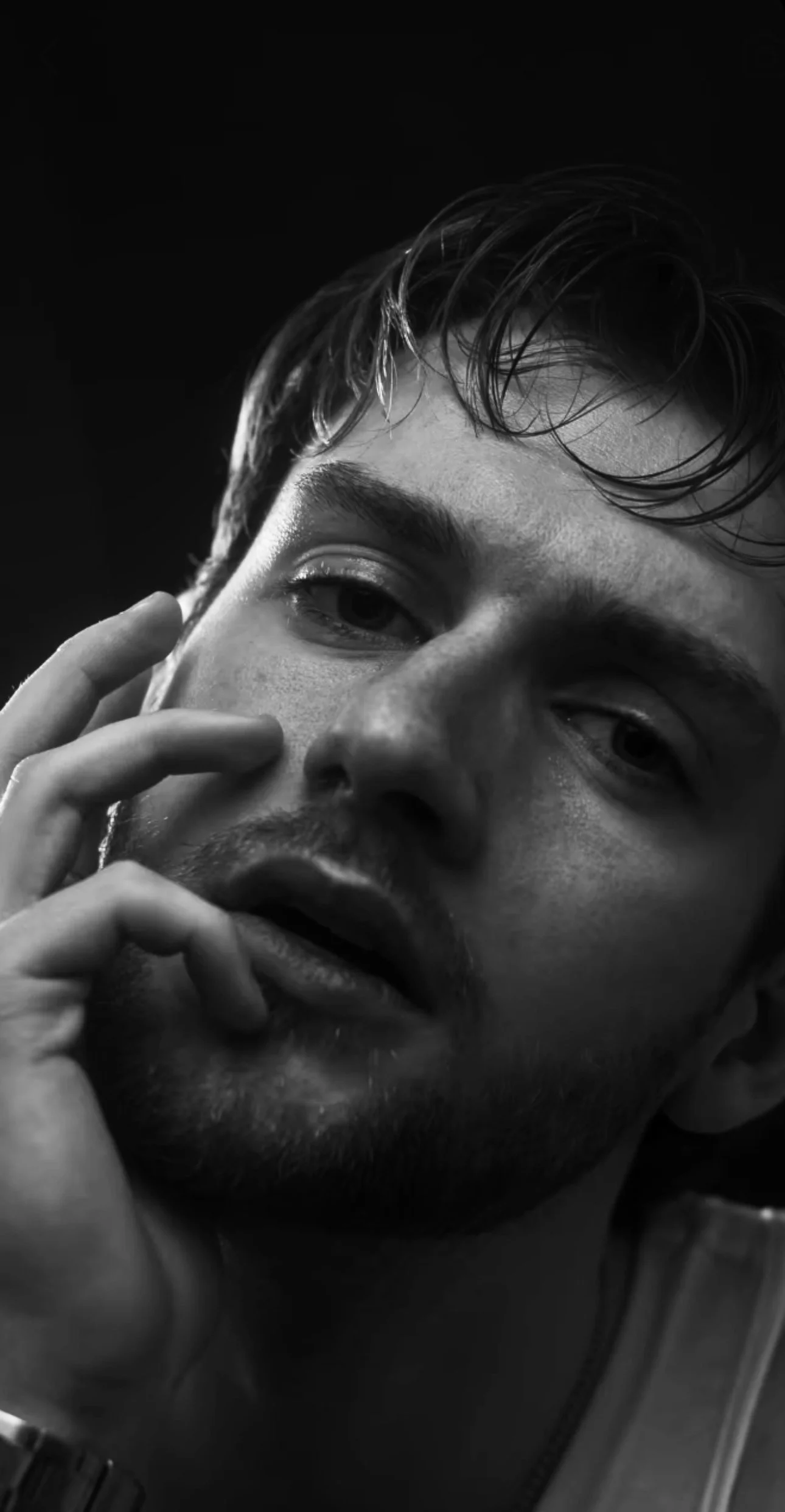 Close-up black and white portrait of a young man, Lyall Hardy Grey, with messy hair, facial hair, and a contemplative expression, touching his face with two fingers.