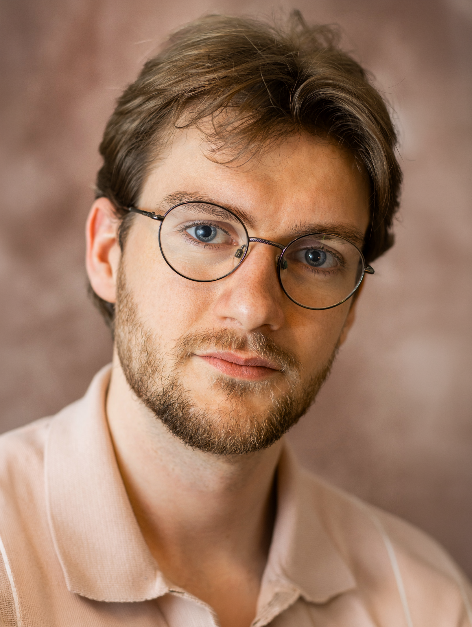 Close-up portrait of Lyall Hardy Grey with brown hair, blue eyes, glasses, light skin, and a beard, wearing a beige jacket and a cream shirt, against a blurred brown background.