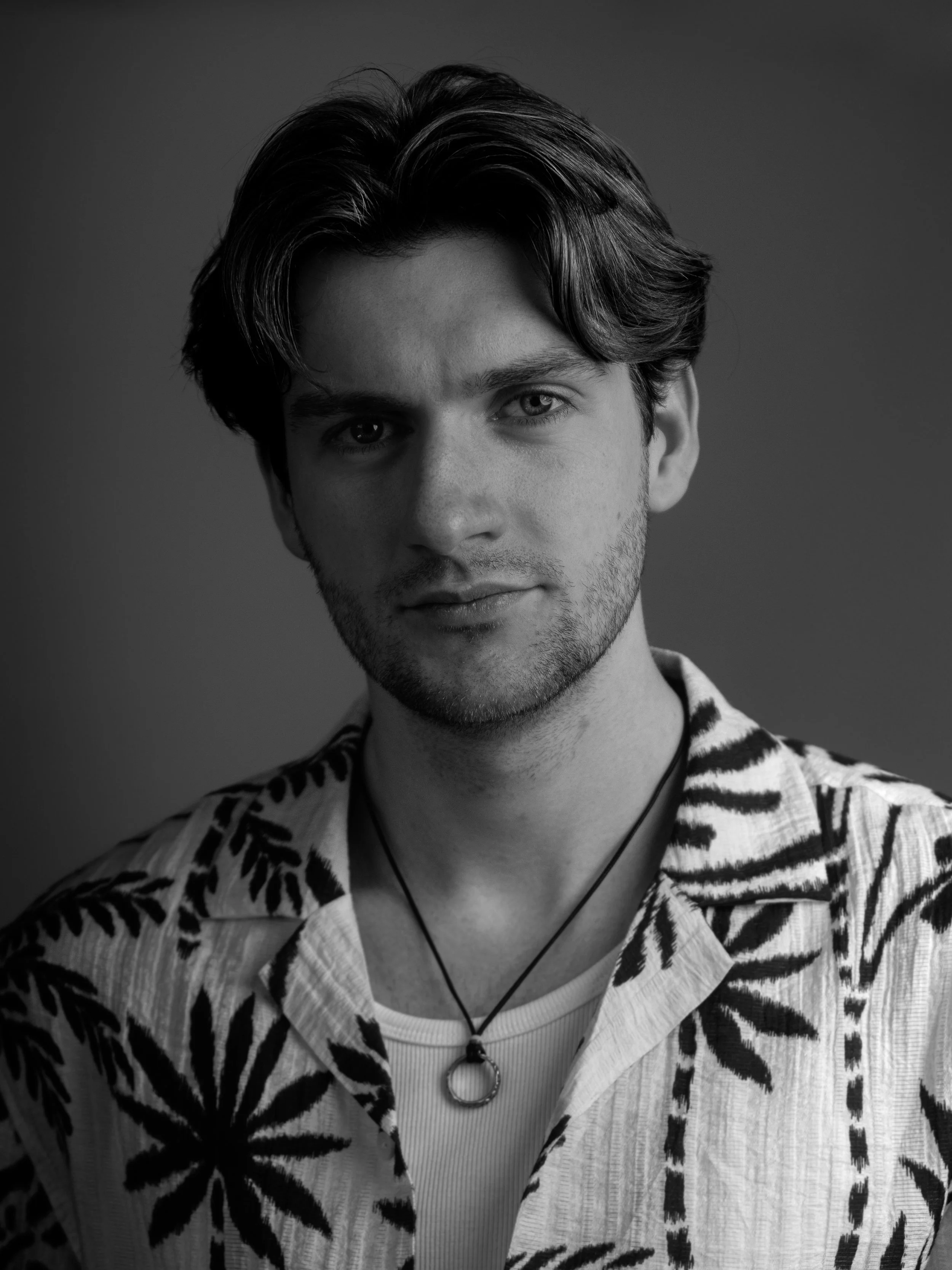 Black and white portrait of a young man, Lyall Hardy Grey, with wavy hair, wearing a zebra-patterned jacket over a light-colored hoodie, and a necklace with a circular pendant.