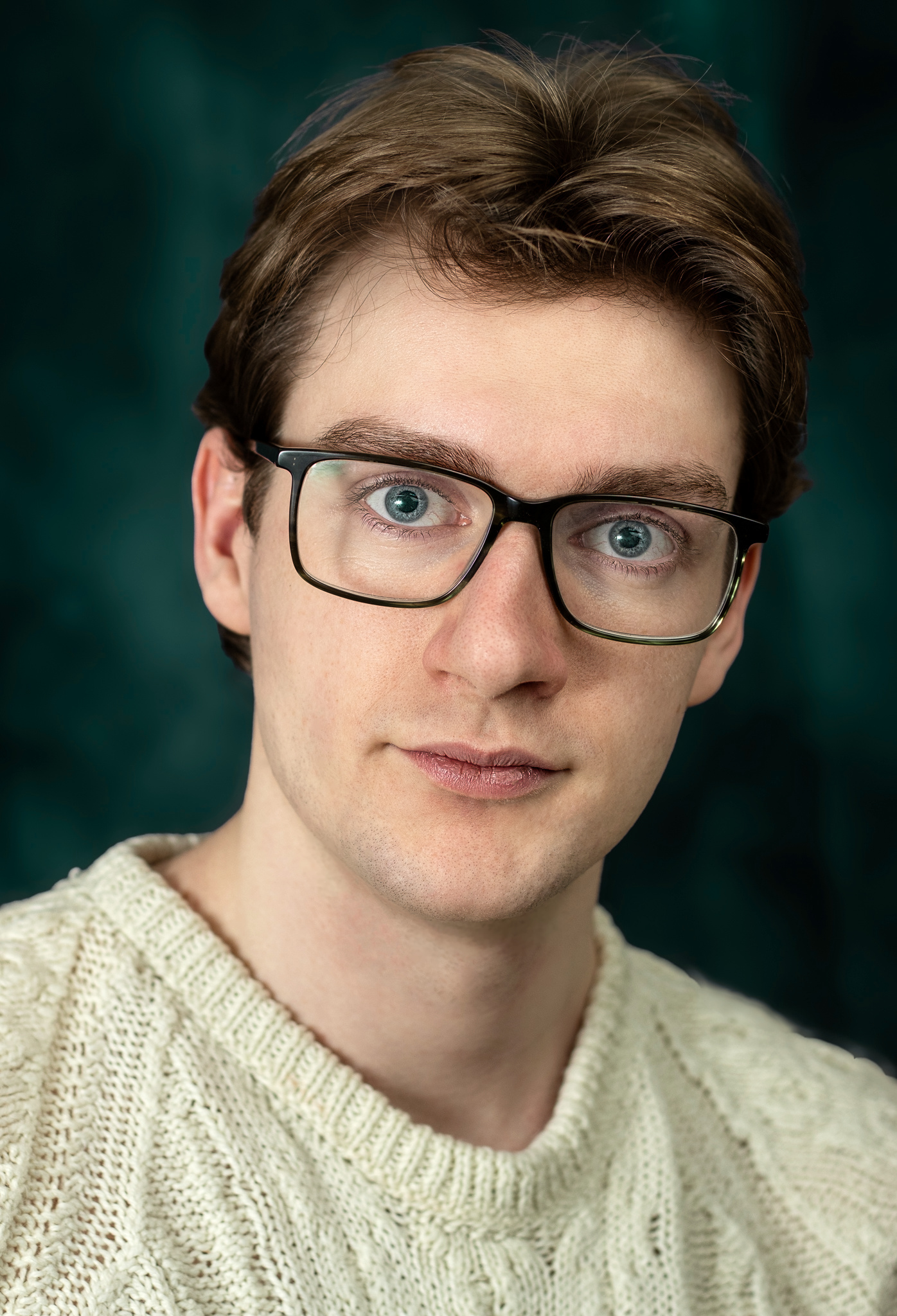 Close-up of Lyall Hardy Grey. A young person wearing glasses and a cream-colored knitted sweater, with short brown hair and blue eyes, looking slightly to the side.