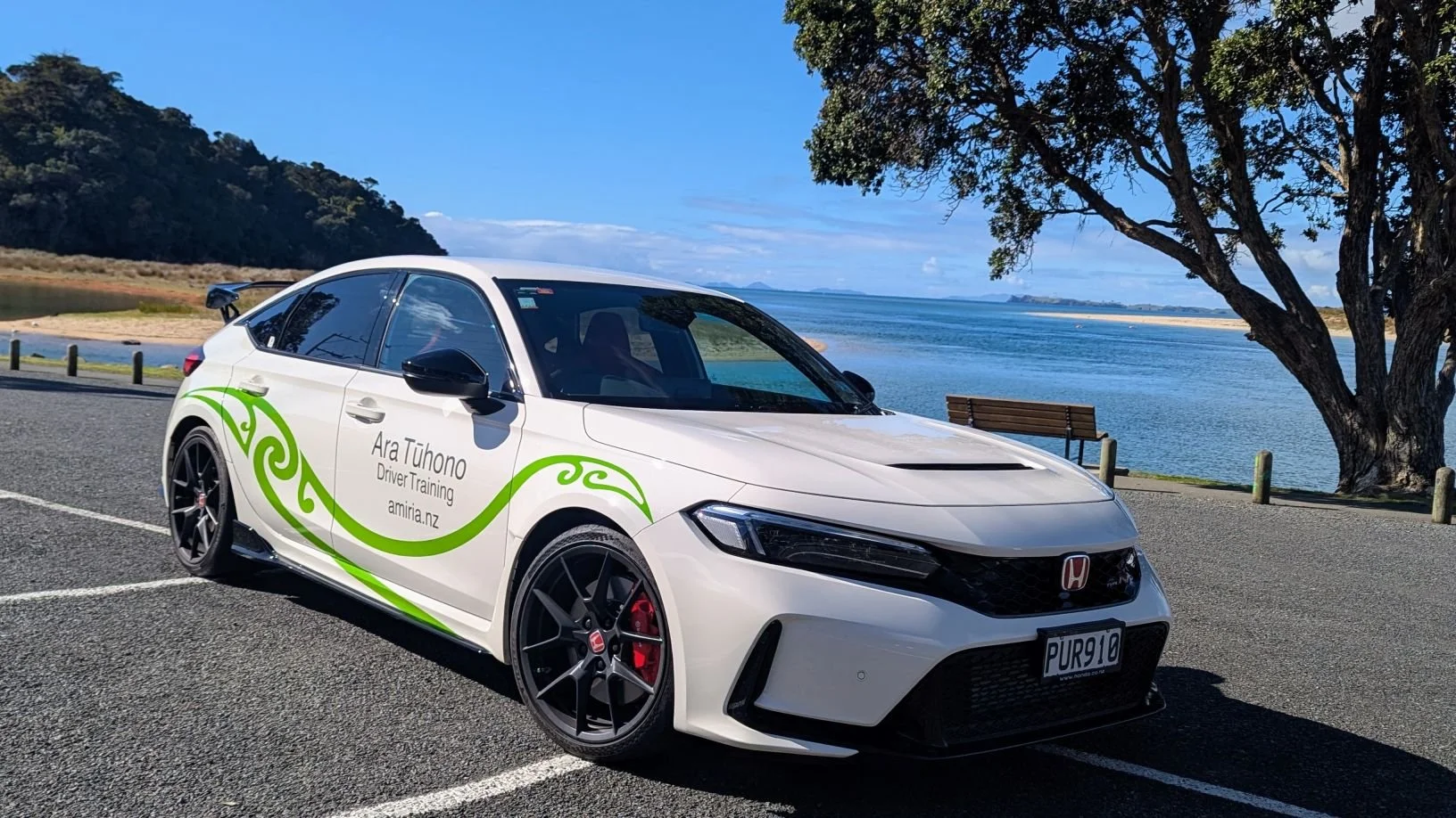 White Honda Civic parked by a lakeside, with green Maori-style decals, a tree, a bench, and a landscape with water, hills, and blue sky in the background.