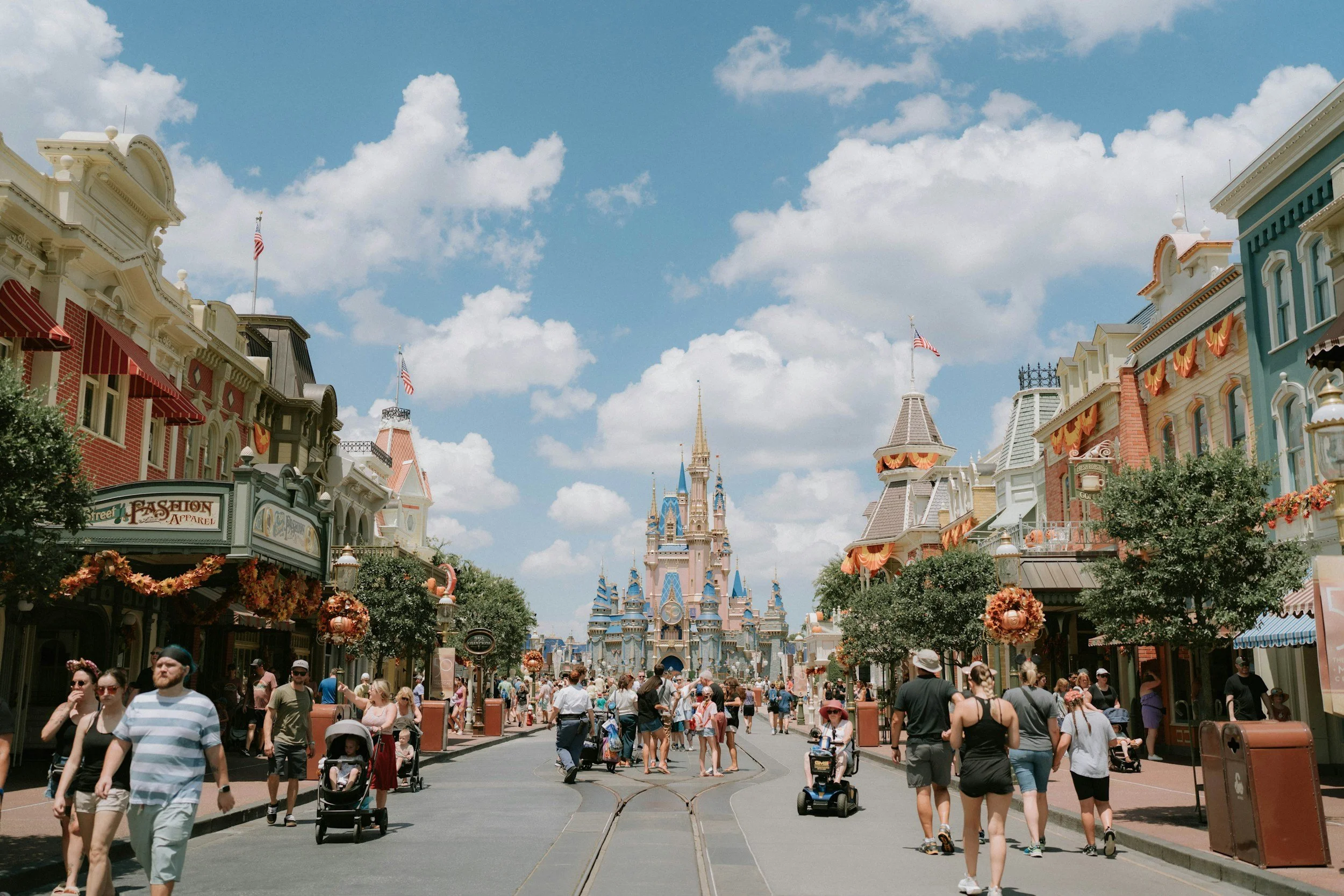 People walking and strolling through a ride at Disneyland, with Sleeping Beauty Castle visible in the background under a partly cloudy sky. The street is decorated with fall or Halloween themed decorations.