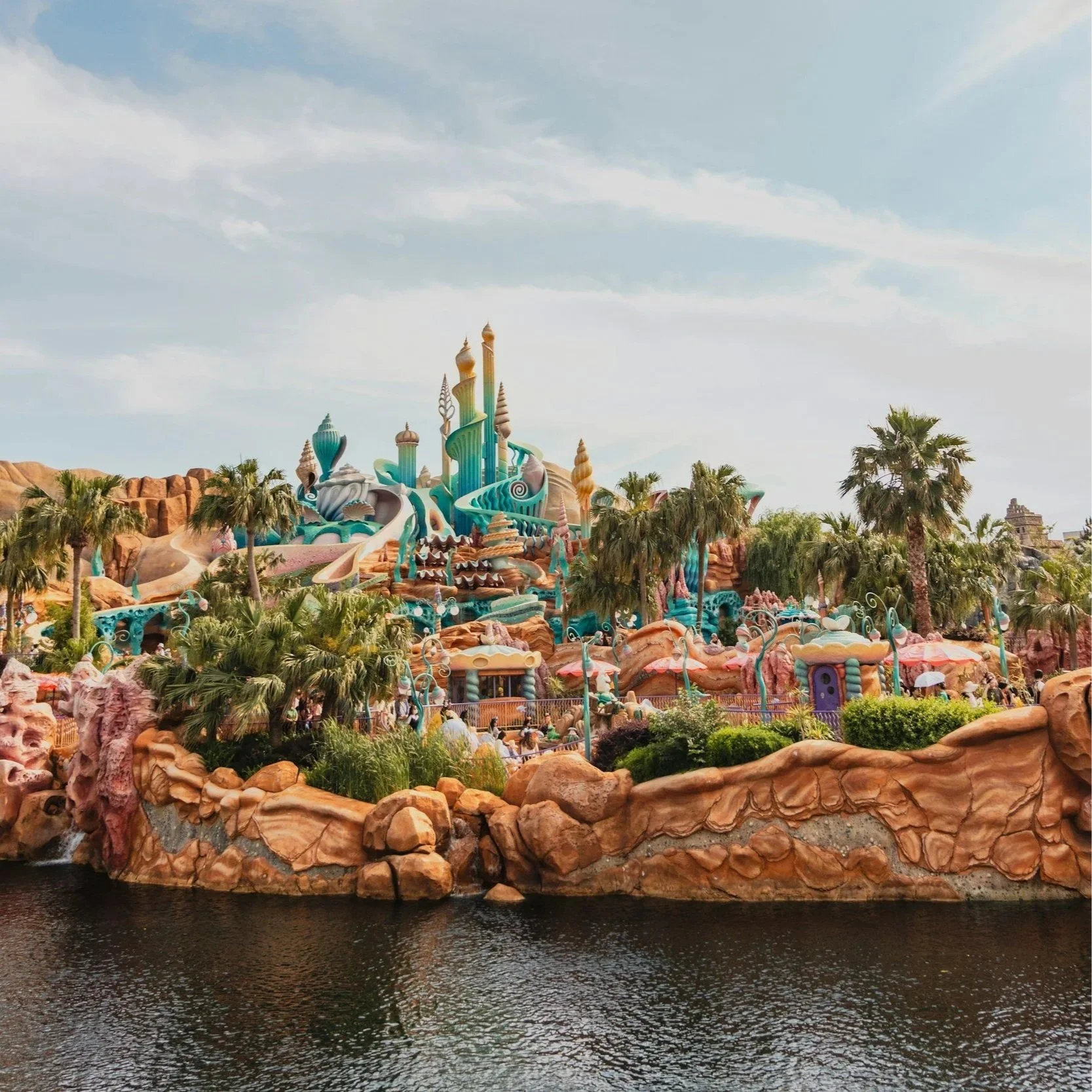 Colorful, whimsical castle with pink, blue, turquoise, and beige spires, surrounded by lush palm trees, with a rocky foreground and water at the bottom, under a partly cloudy sky.