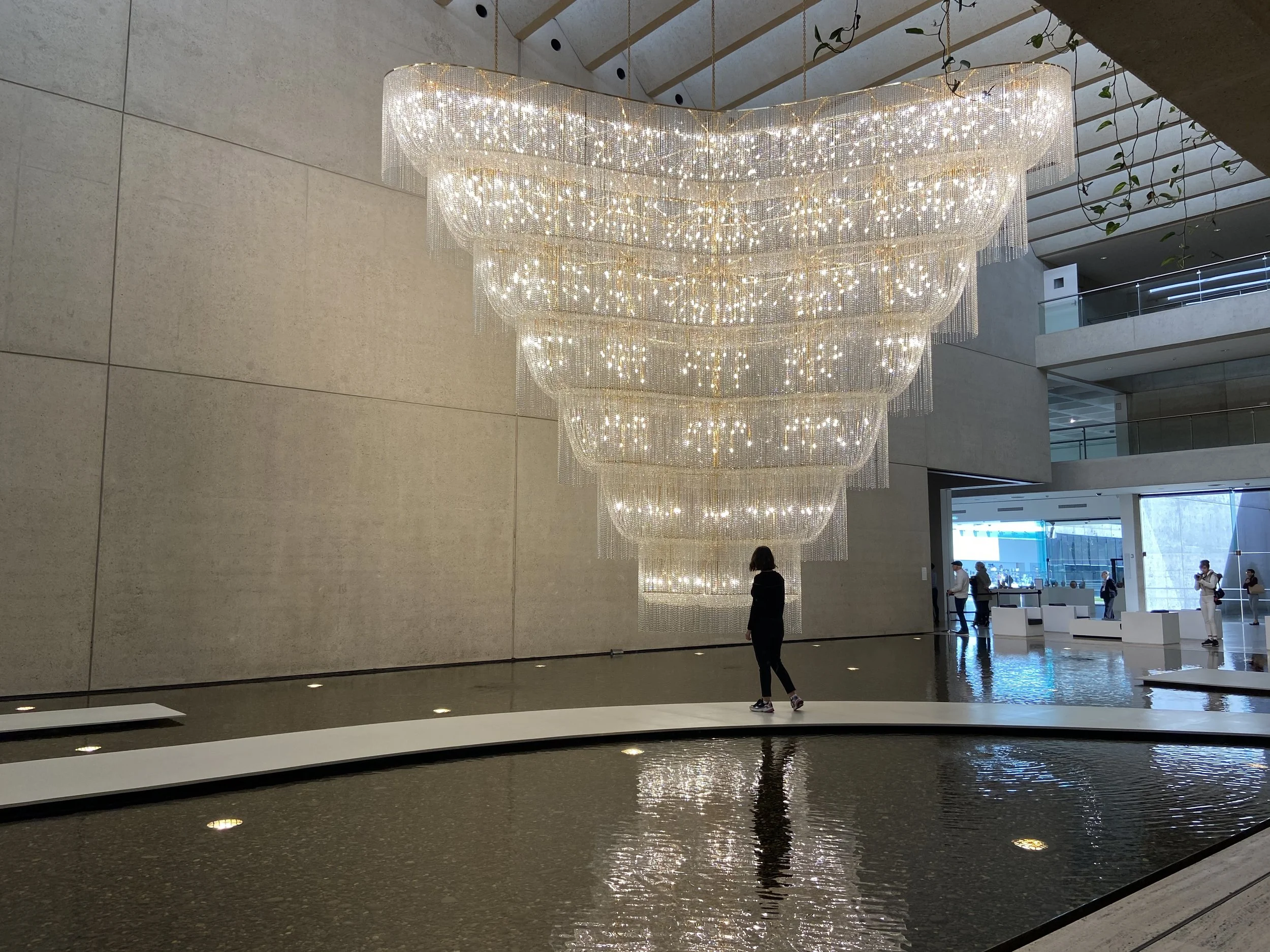 Large, ornate chandelier with multiple tiers of glowing crystals hanging inside a modern building with beige tiled walls and a water feature in the foreground.