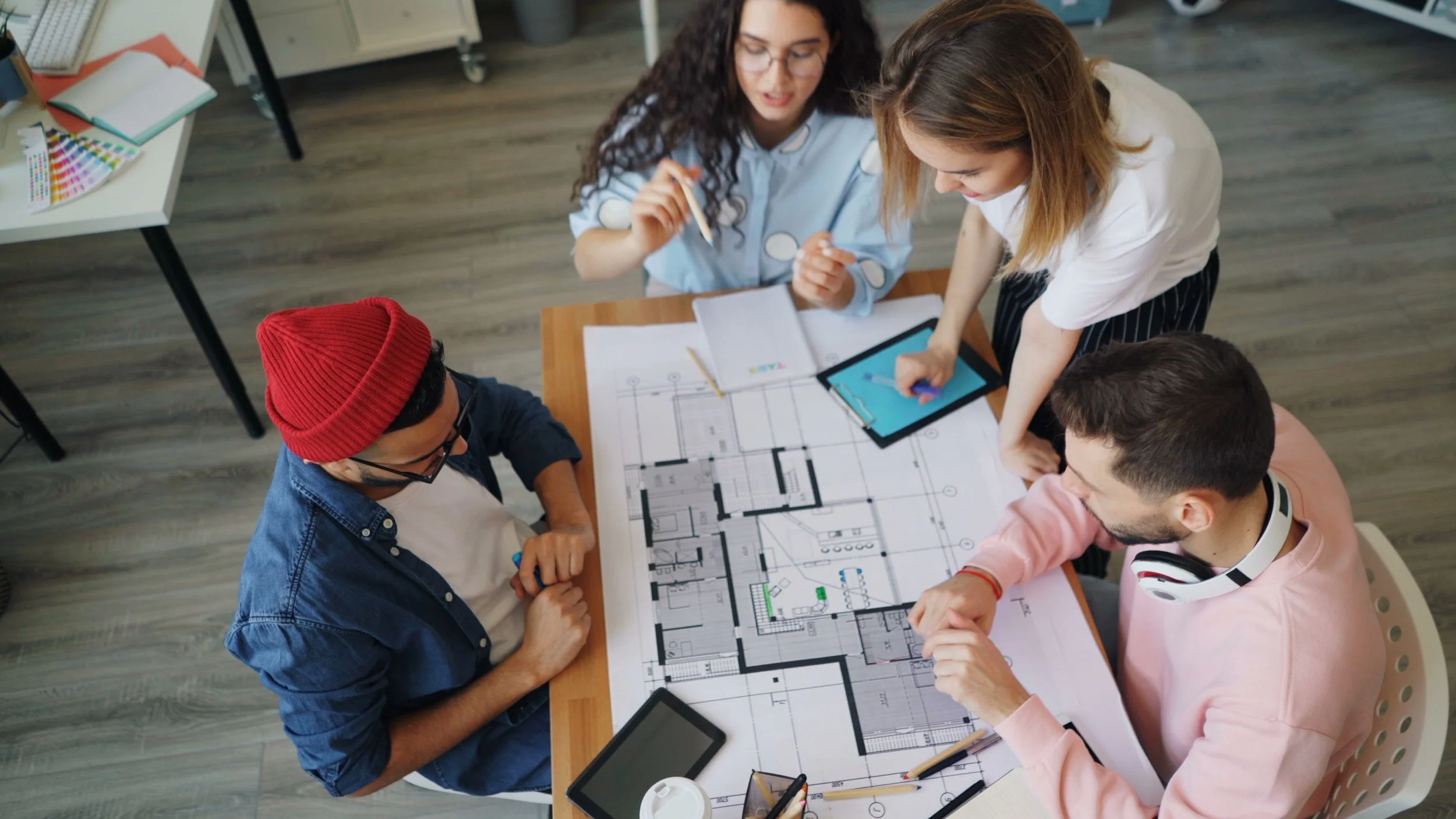 Group of four young adults collaborating on building floor plans at a desk, with various tools and a tablet.
