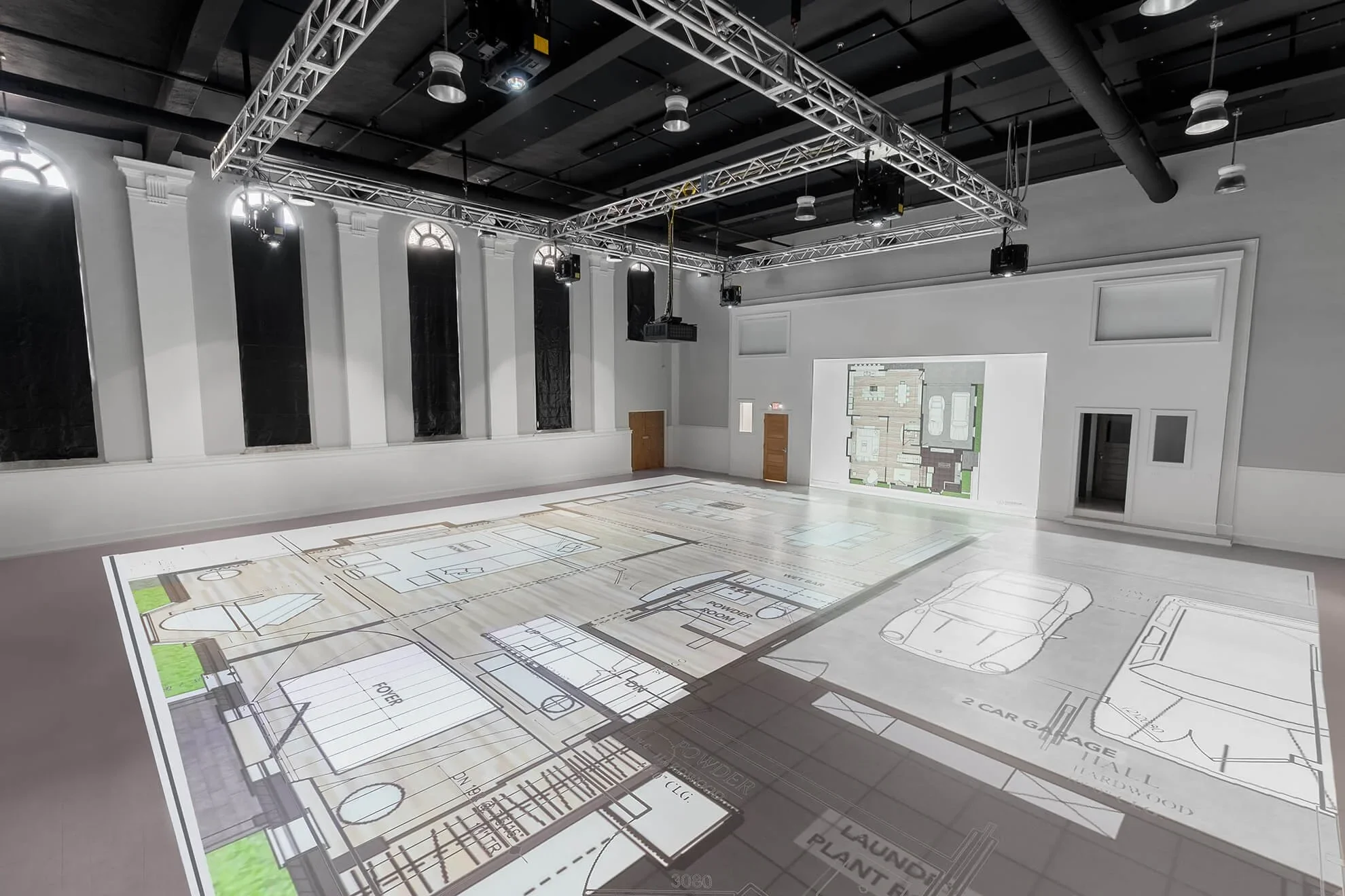 An indoor event space with a metal truss ceiling system, theatrical lighting, and wall-mounted speakers. The white wall has tall narrow arched windows, and a projection shows a floor plan of a building with labeled rooms and garage spaces.