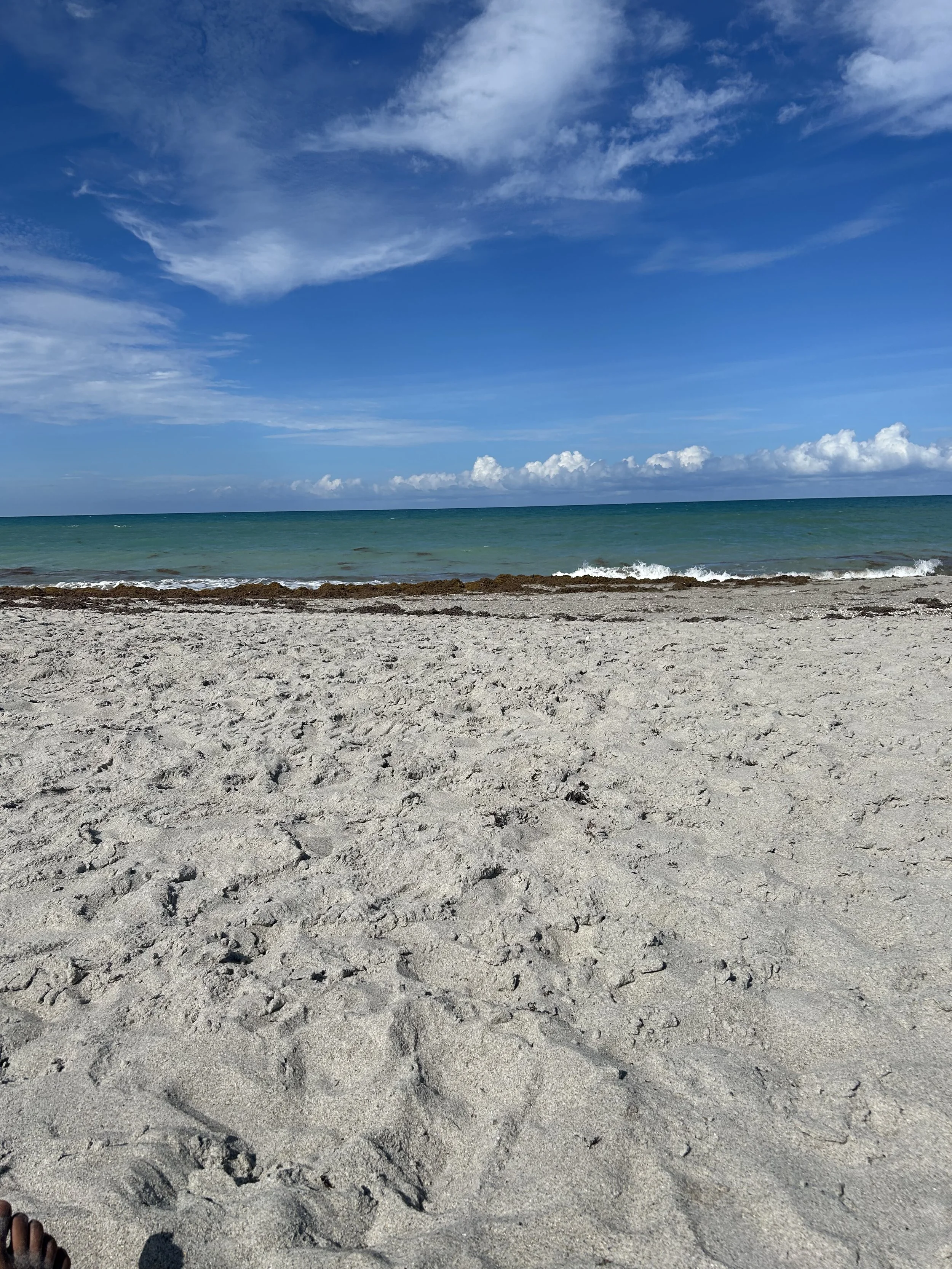A sandy beach with footprints leading into the distance, calm seawater, and a partly cloudy blue sky.