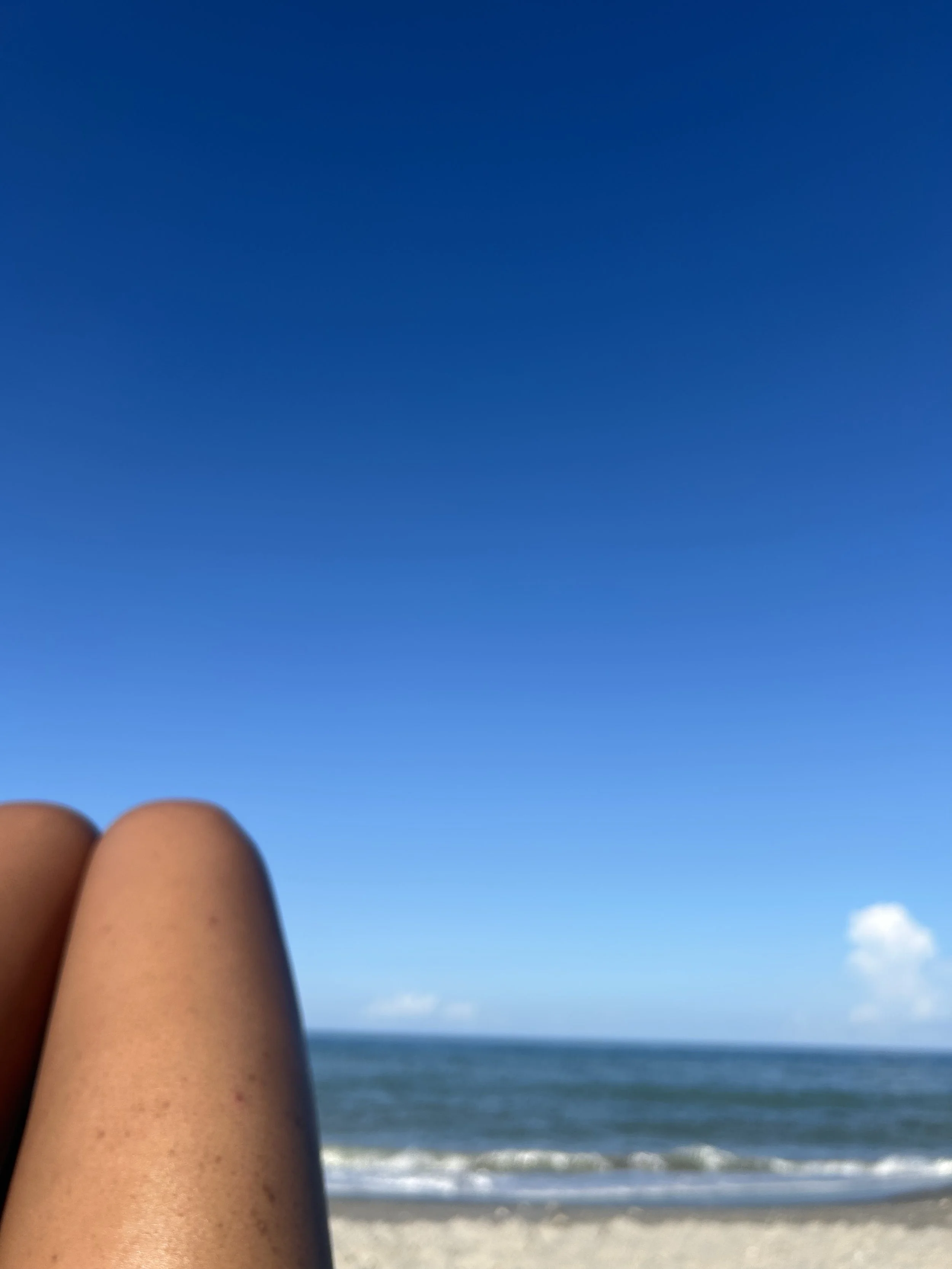 Close-up of tan legs and feet with toes, at the beach, with ocean, sandy shore, blue sky, and a small white cloud in the background.