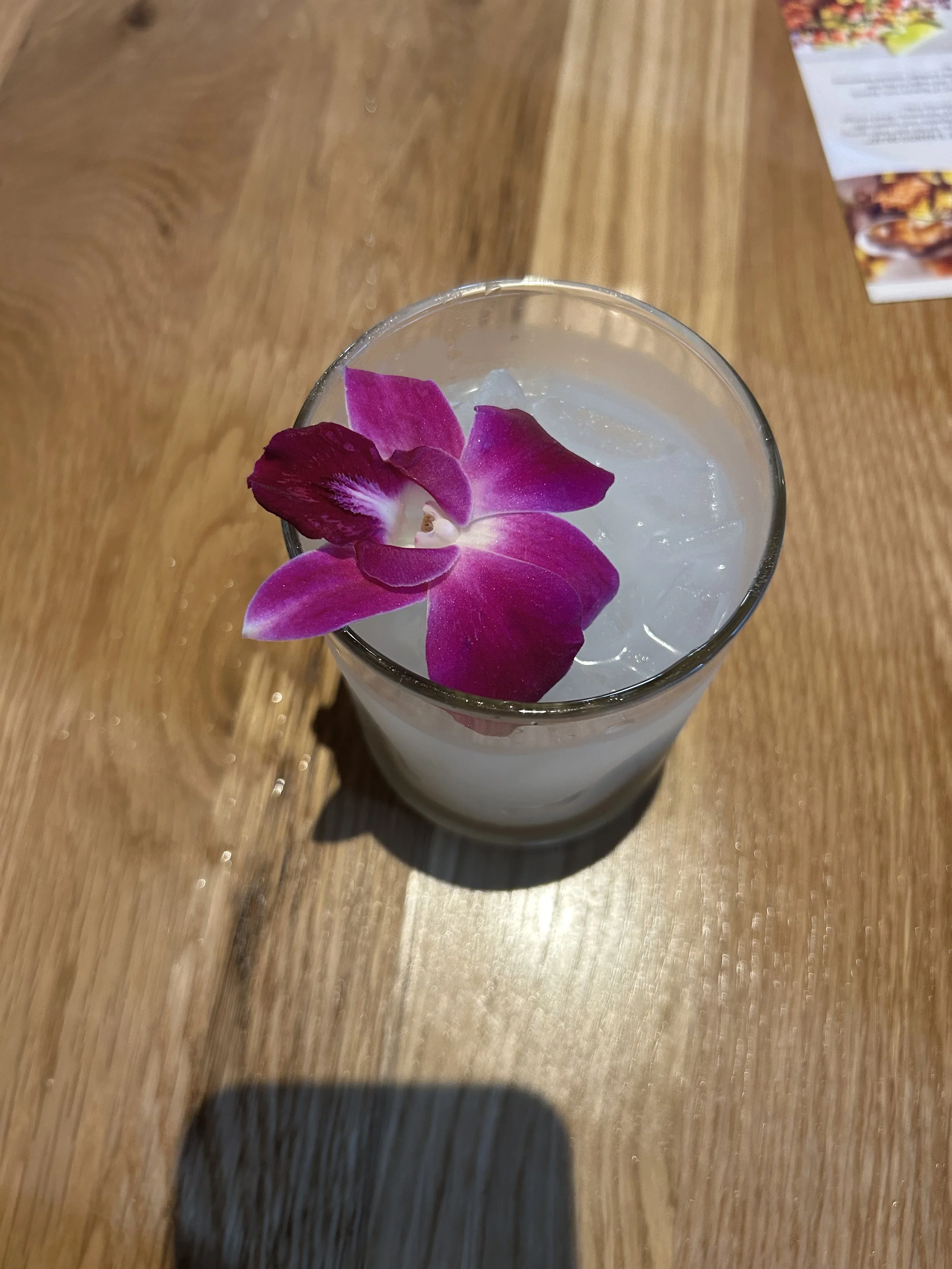 A tall glass with a purple orchid flower on the rim, filled with a clear beverage and ice cubes on a wooden table.