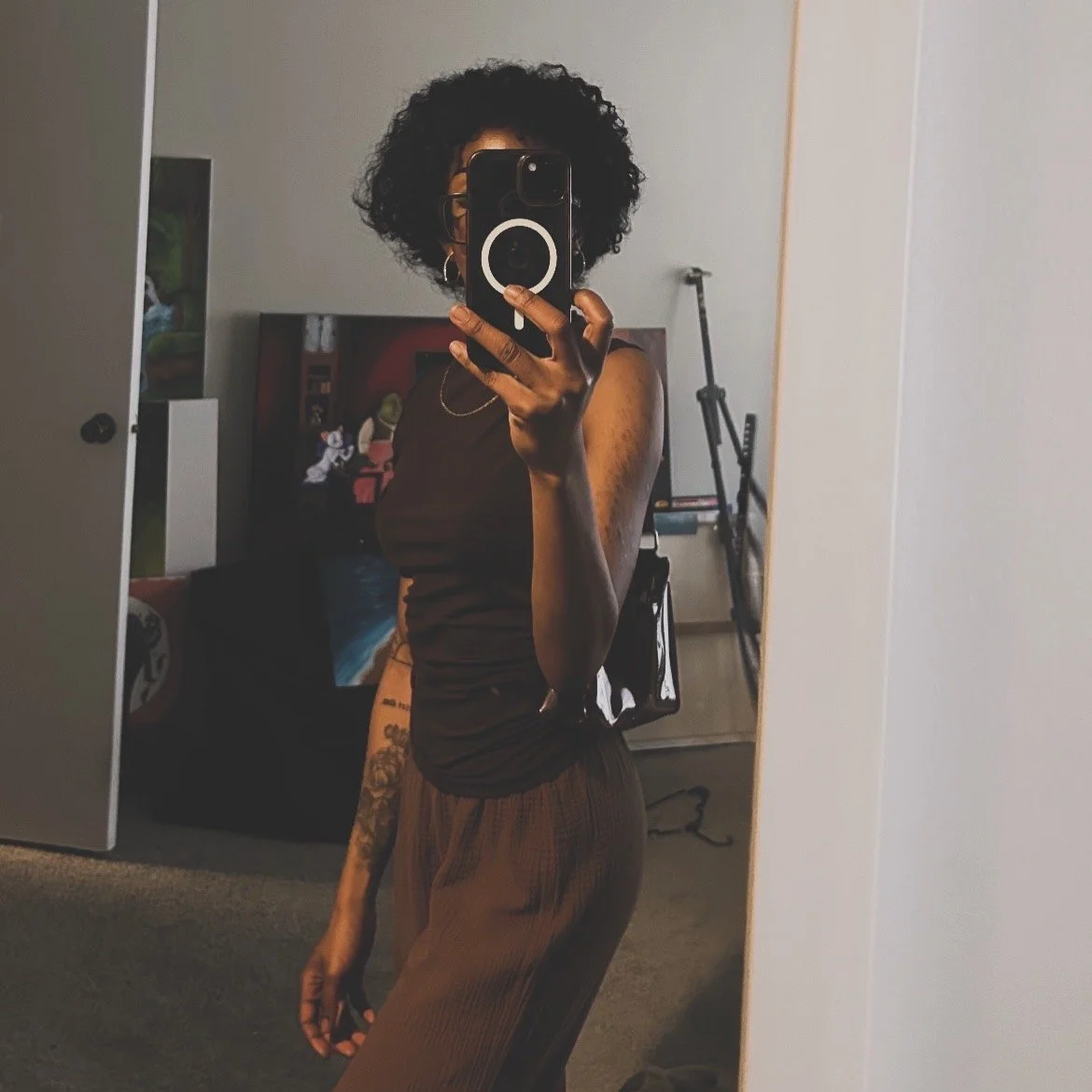 A woman with curly hair taking a mirror selfie in a bedroom, wearing a black sleeveless top and brown pants.