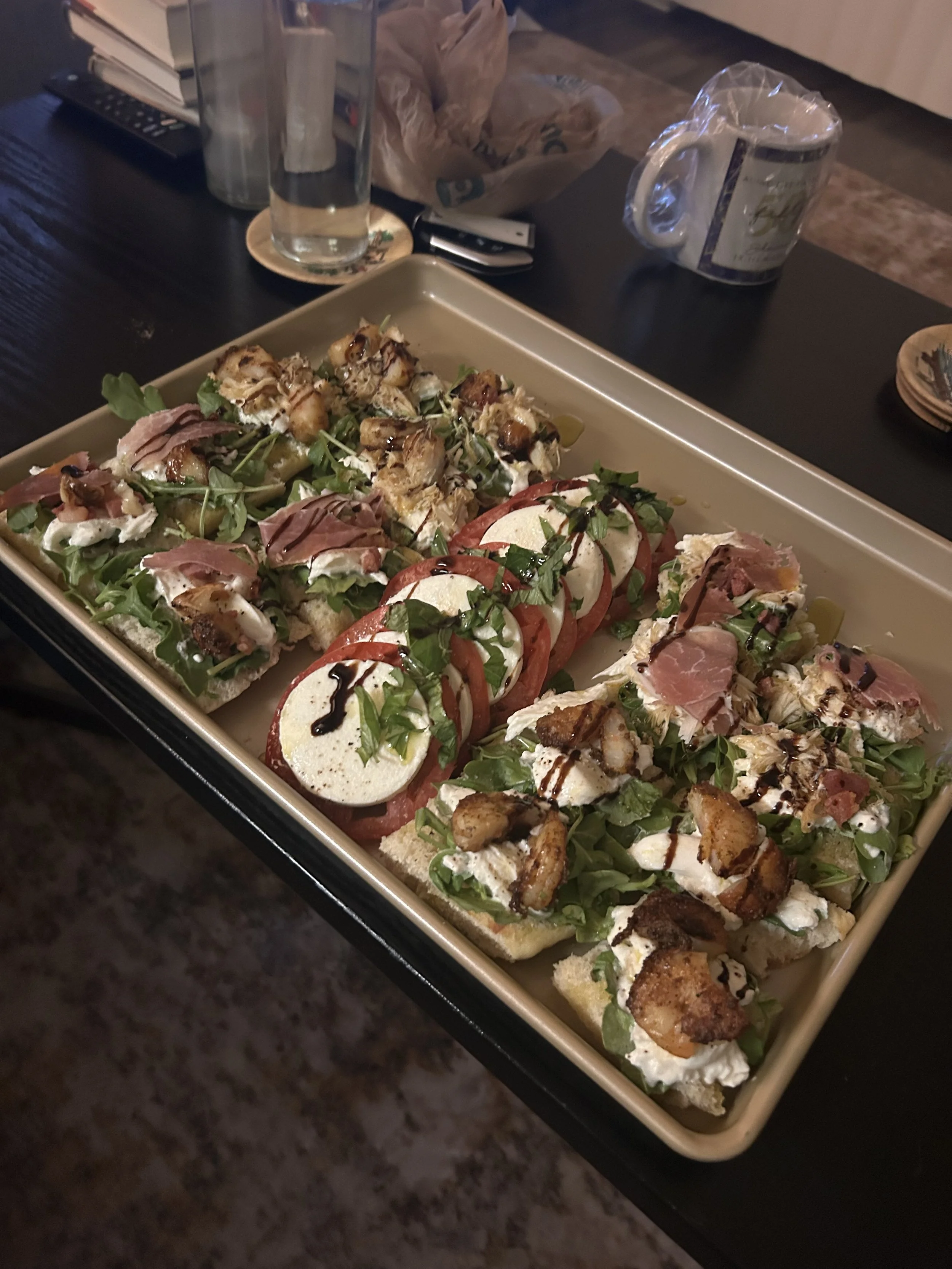 A rectangular serving platter with various types of open-faced sandwiches topped with ingredients such as tomatoes, mozzarella, avocado, meats, greens, and sauces, on a dark table.