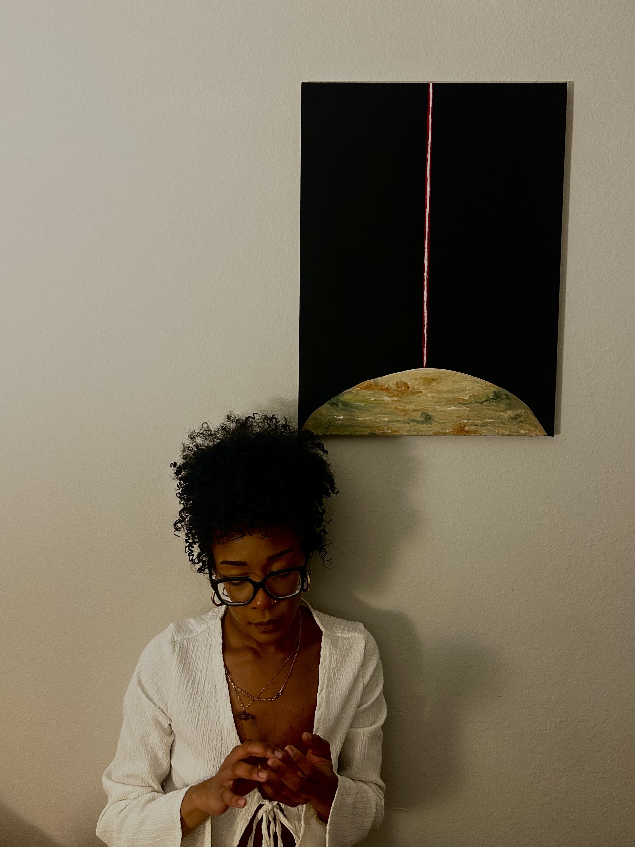 A woman with curly black hair and glasses, wearing a white sweater, standing against a beige wall and looking down at her phone.