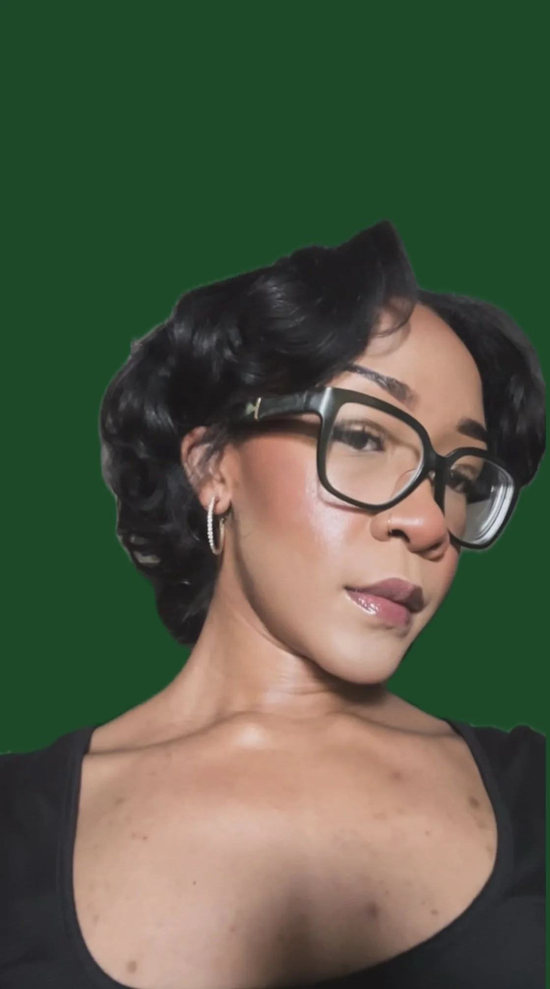 Close-up of a woman with vintage black hair, wearing large dark green glasses, hoop earrings, and a black top, against a dark green background.