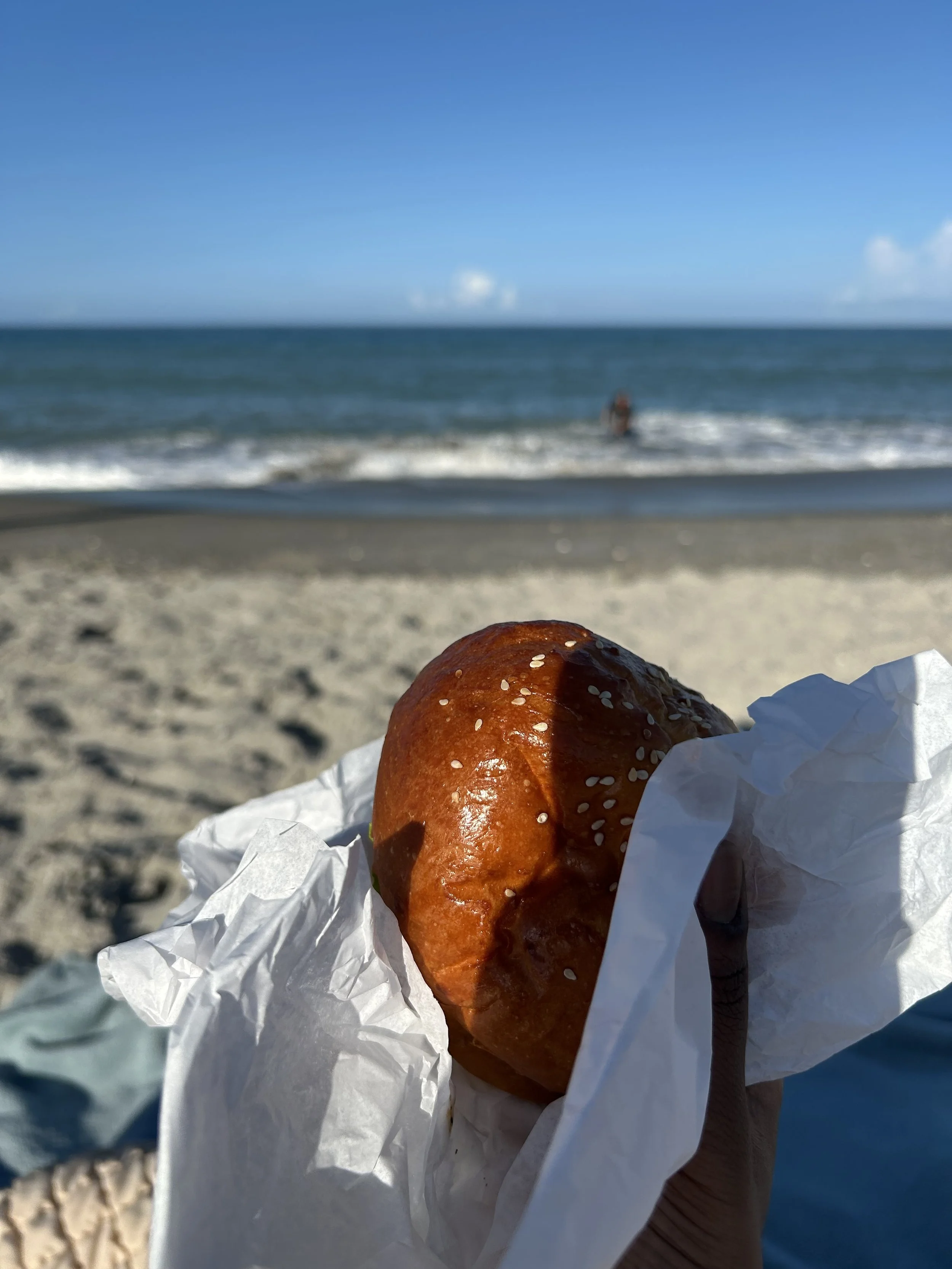 A hand holding a glazed hamburger bun sandwich wrapped in white paper, with the beach and ocean in the background under a clear blue sky.