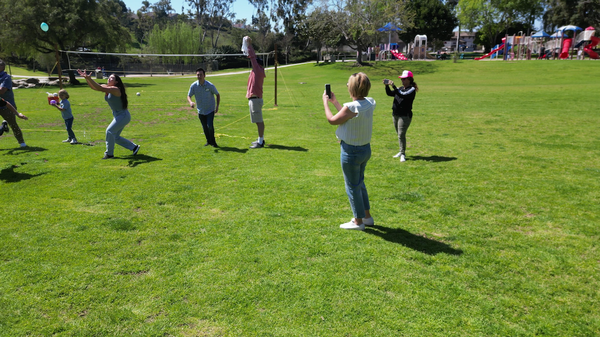 People playing volleyball outdoors on a grassy field with playground equipment in the background.