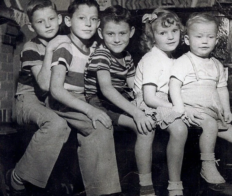 Five children sitting together on a bench, posing for a black-and-white photograph. They are wearing casual clothing and appear to be in a vintage setting.