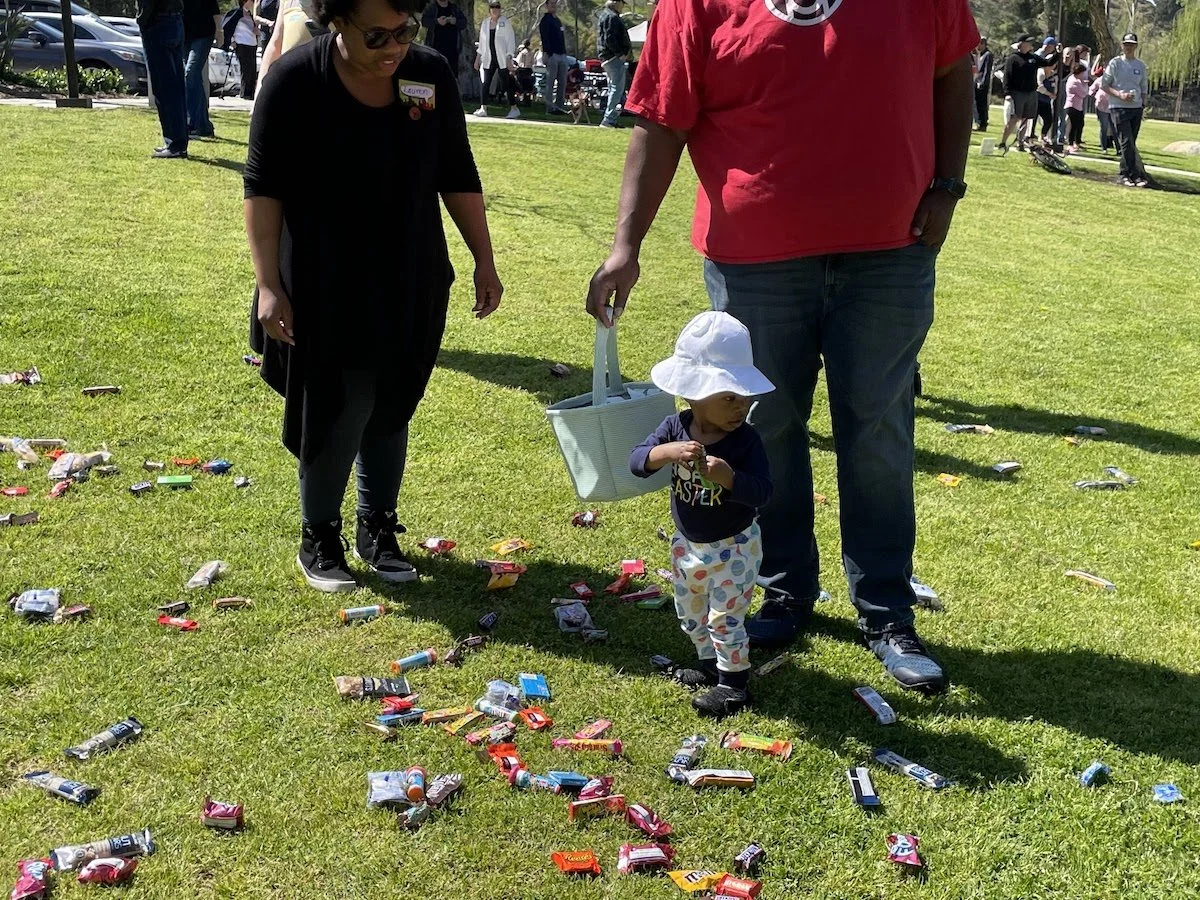 A child with a white sun hat and dark shirt picking up candy from a grassy area, with an adult holding a shopping bag, and a woman standing nearby, while many people gather in the background on a sunny day.