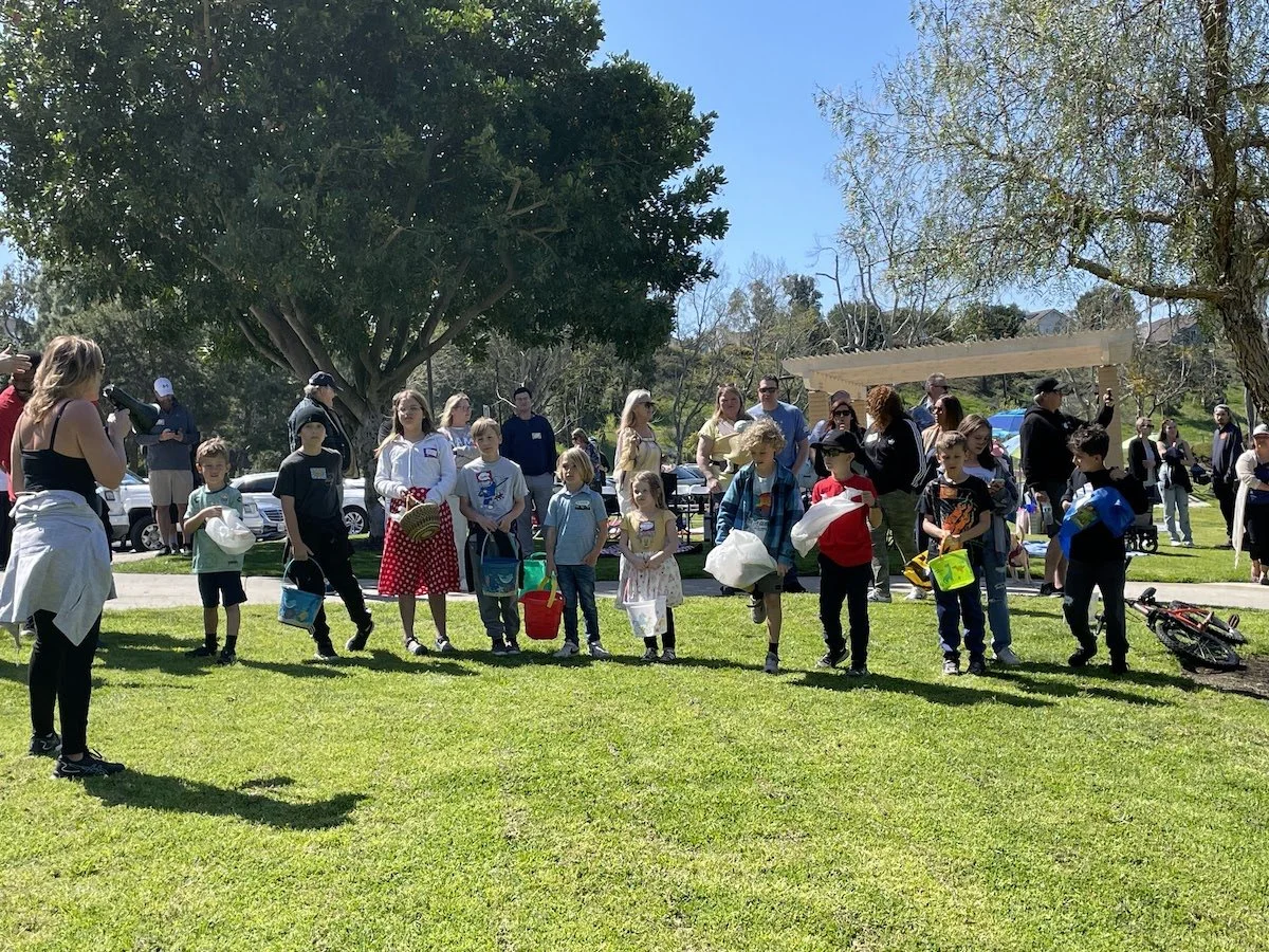 Children and adults standing in line outdoors on a sunny day, some with buckets and bags, possibly waiting for an event or activity in a park.
