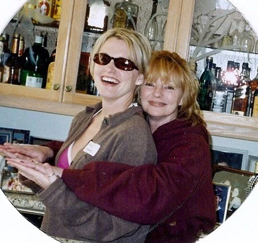Two women smiling and hugging in a kitchen, with glass cabinets and bottles in the background.