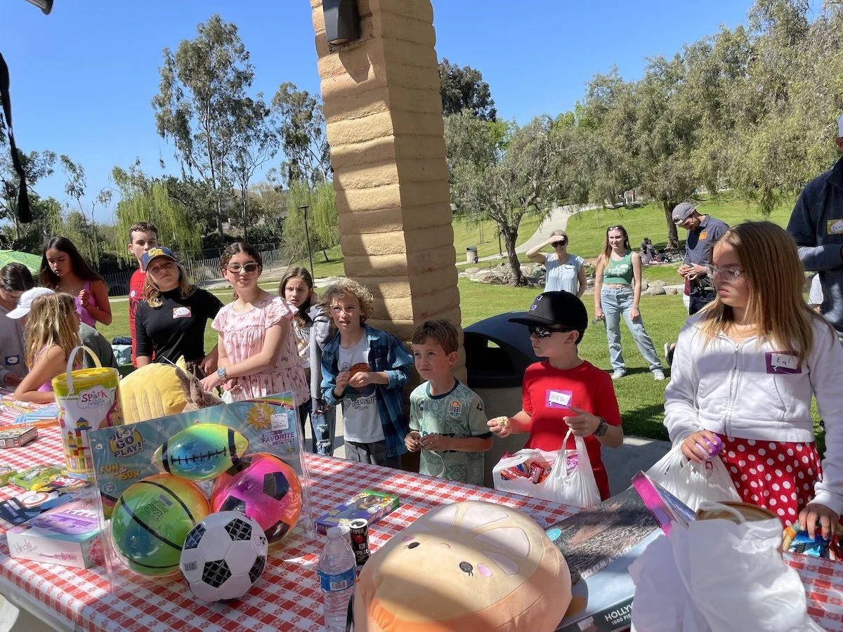 Children and adults gathered outdoors around a table filled with toys at a daytime event in a park with trees and a clear blue sky.