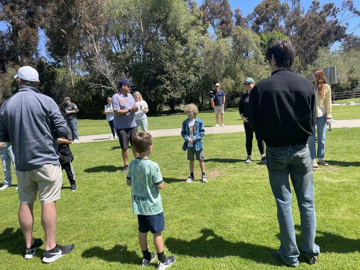 Group of children and adults gathered on a grassy park, listening to a man in a blue shirt speaking. Some people are taking photos or videos. There are trees in the background and a sunny sky.