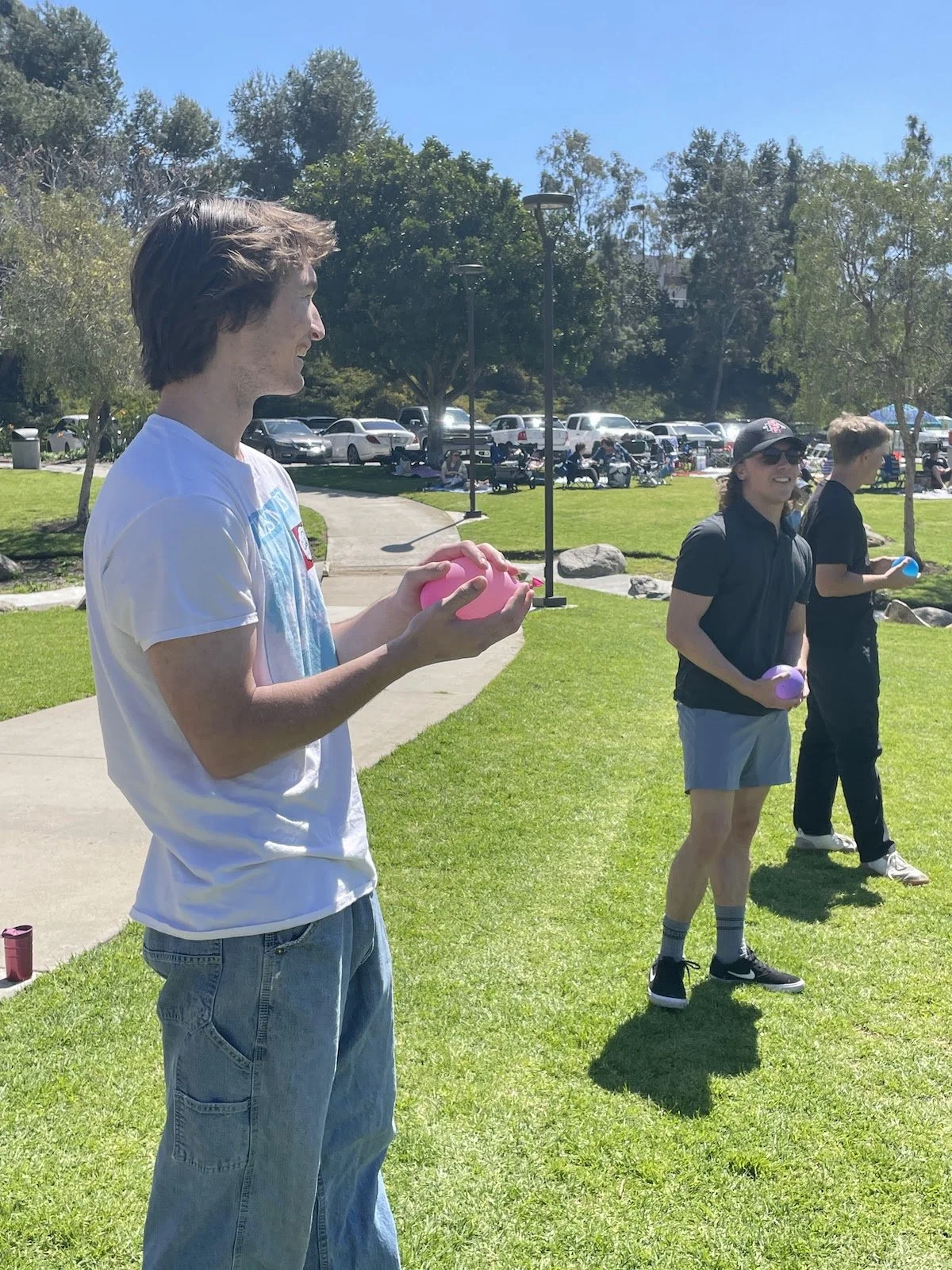 Three young people standing on a grassy field, each holding a small colorful balloon, with a park and parking lot in the background on a sunny day.