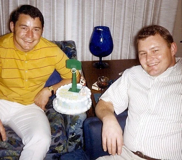 Two men sitting on a sofa celebrating birthday, with a cake and a blue vase on the table.