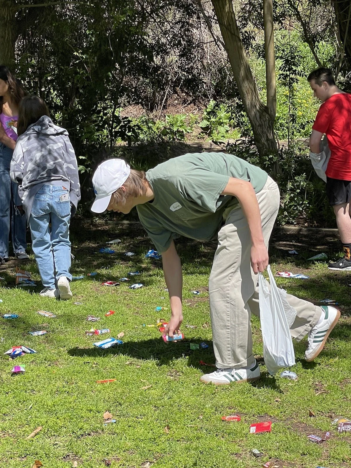 People participating in a cleanup activity outdoors, collecting trash on the grass near trees.