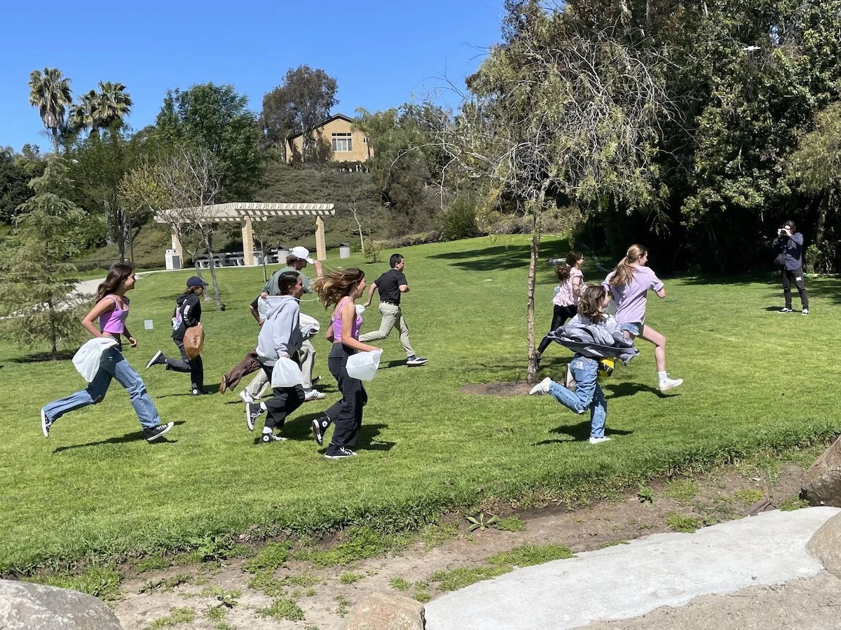 Group of children running outdoors on a grassy lawn with trees, under a clear blue sky, with an adult taking photos in the background.