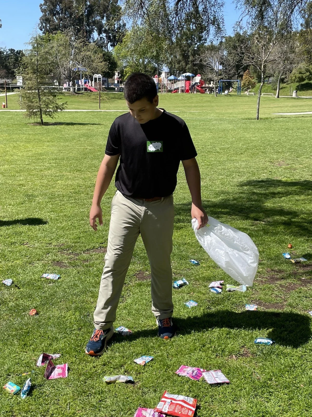 A young boy standing on a grassy field holding a plastic trash bag and picking up litter. The field is scattered with snack wrappers and food packaging. In the background, there is a playground with slides and trees.