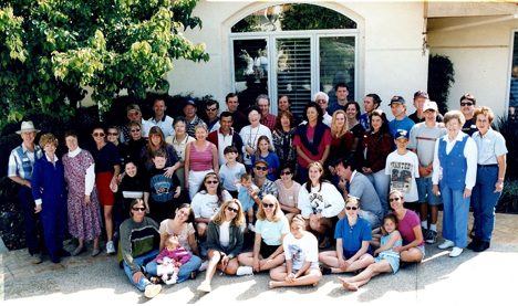 A large group of people gathered outside a house in front of a window and door, posing for a group photo. The group includes men, women, and children of various ages, with some wearing casual clothing, hats, and sunglasses.