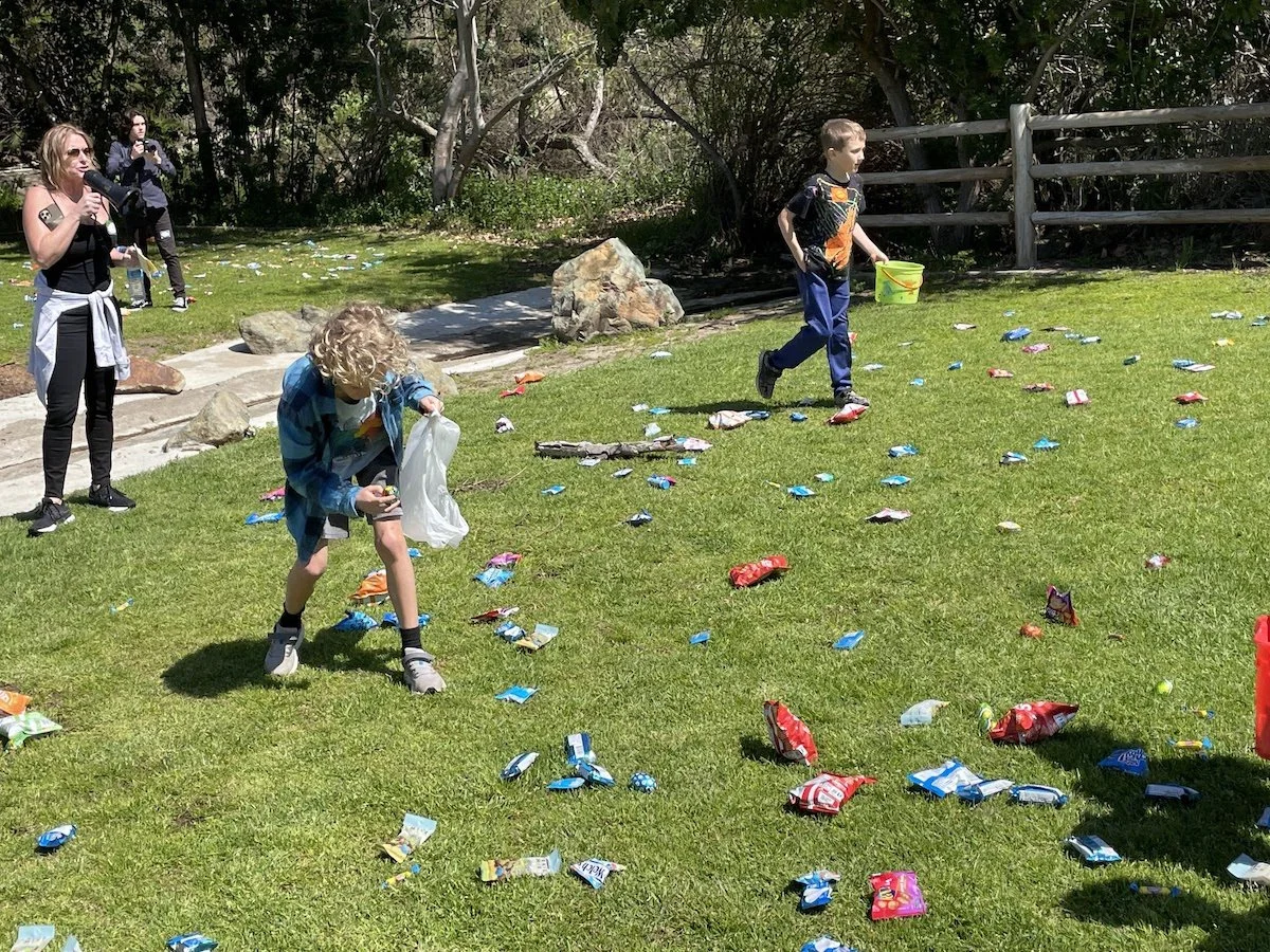 Children participating in an outdoor game of catching candy candies scattered on a grassy field, with people observing nearby.
