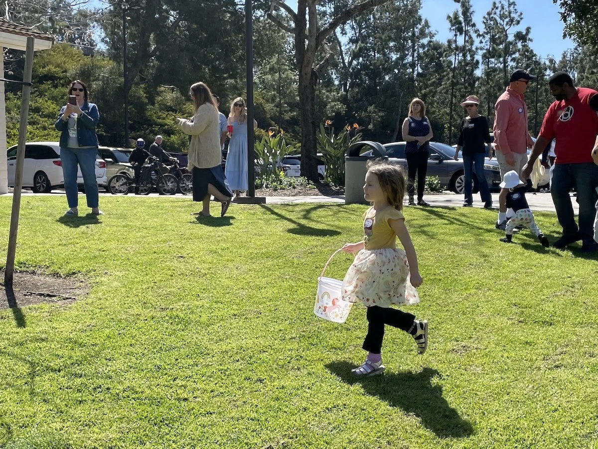 A young girl with a yellow shirt and floral skirt walking on the grassy park, holding a bucket, while people in the background gather and walk around.
