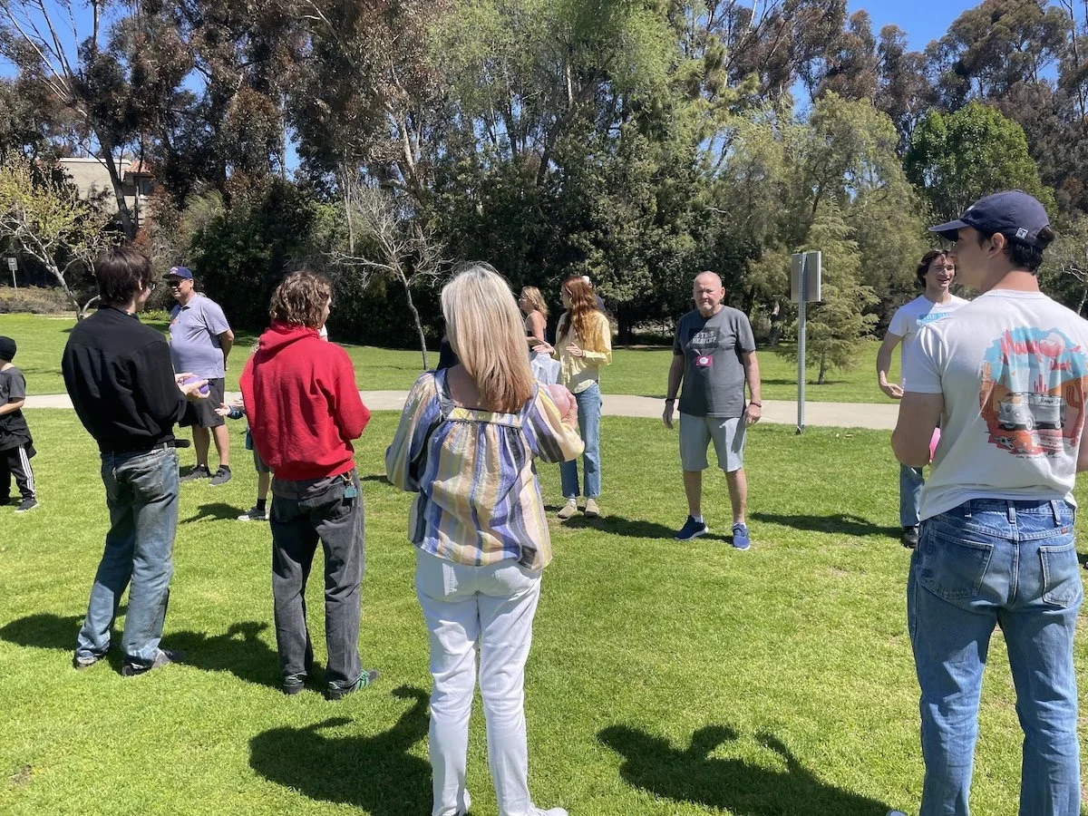 Group of people standing on a grass field outdoors on a sunny day, chatting and socializing.