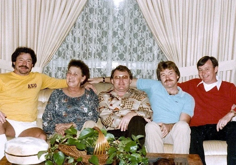 A group of five people sitting on a beige couch in a living room with lace curtains. They are smiling and have their arms around each other, with a coffee table and potted plant in front.