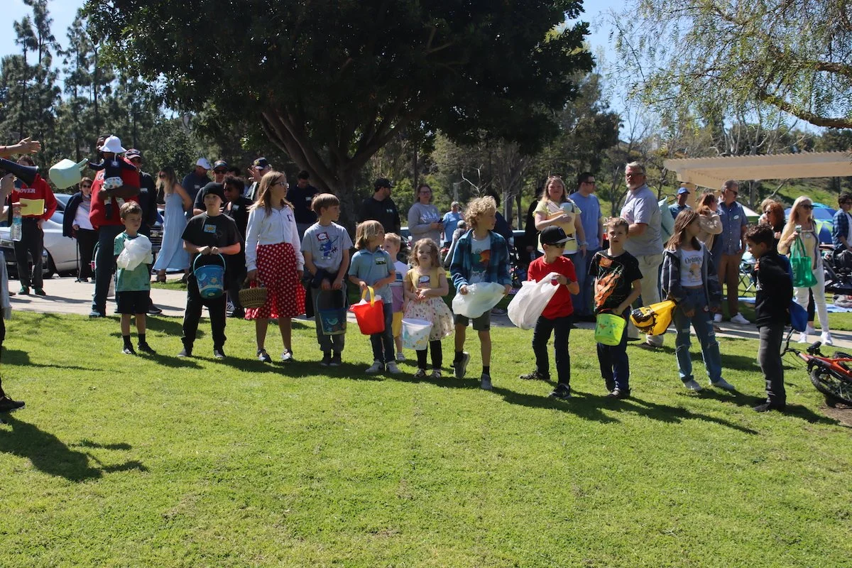 Children standing in a line outdoors with adults in the background, in a park with trees, on a sunny day.