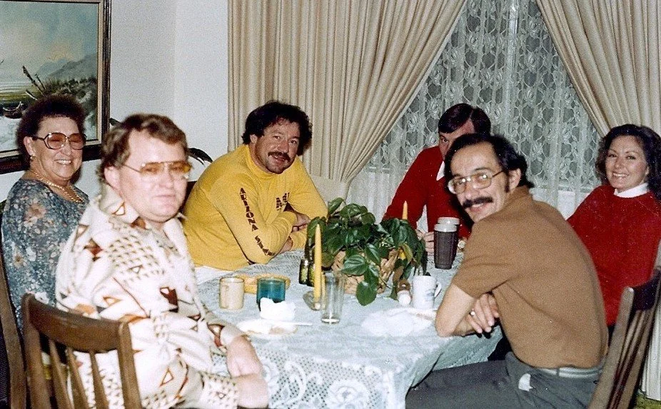 Vintage photograph of six people, three women and three men, gathered around a dining table in a well-lit room with curtains and a landscape painting on the wall.