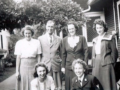 Black and white photo of six women standing and sitting outdoors in front of a house and trees, dressed in vintage clothing.