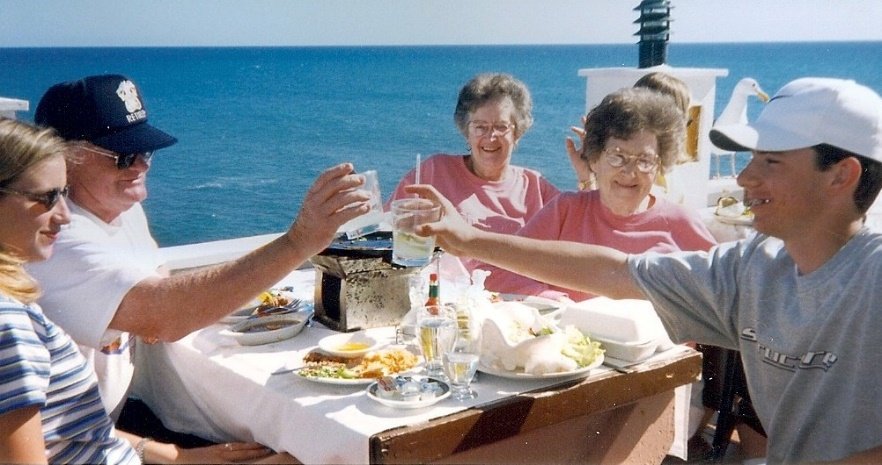 Group of five people, including three women and two men, enjoying a meal outdoors on a boat with the ocean and a lighthouse in the background, raising glasses for a toast.