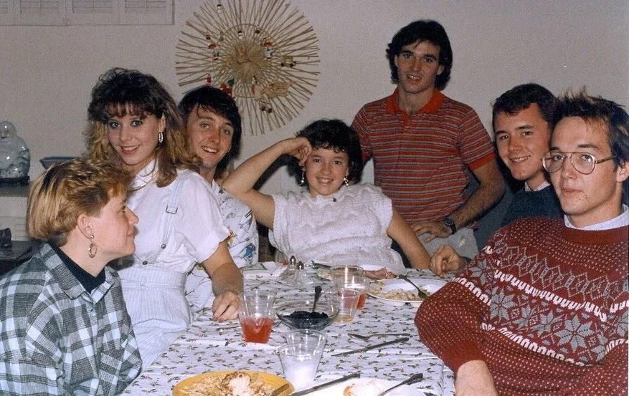 A group of seven young people sitting around a decorated table, enjoying a meal and smiling for the photo indoors.