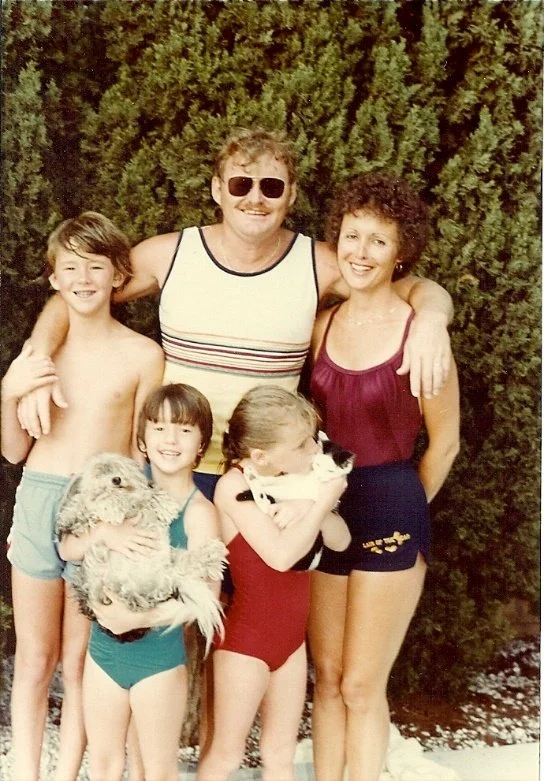 A group of five people and two dogs standing outdoors in front of a green hedge, smiling and posing for a photo.