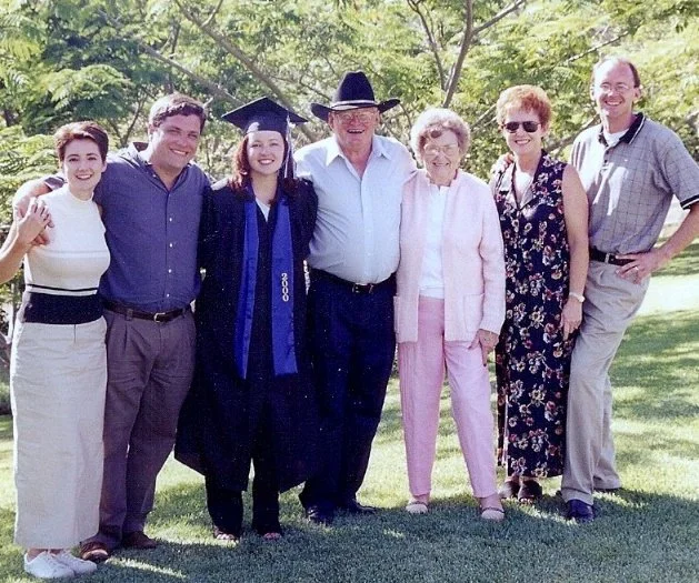 Family group celebrating graduation outdoors; woman in cap and gown in the center.